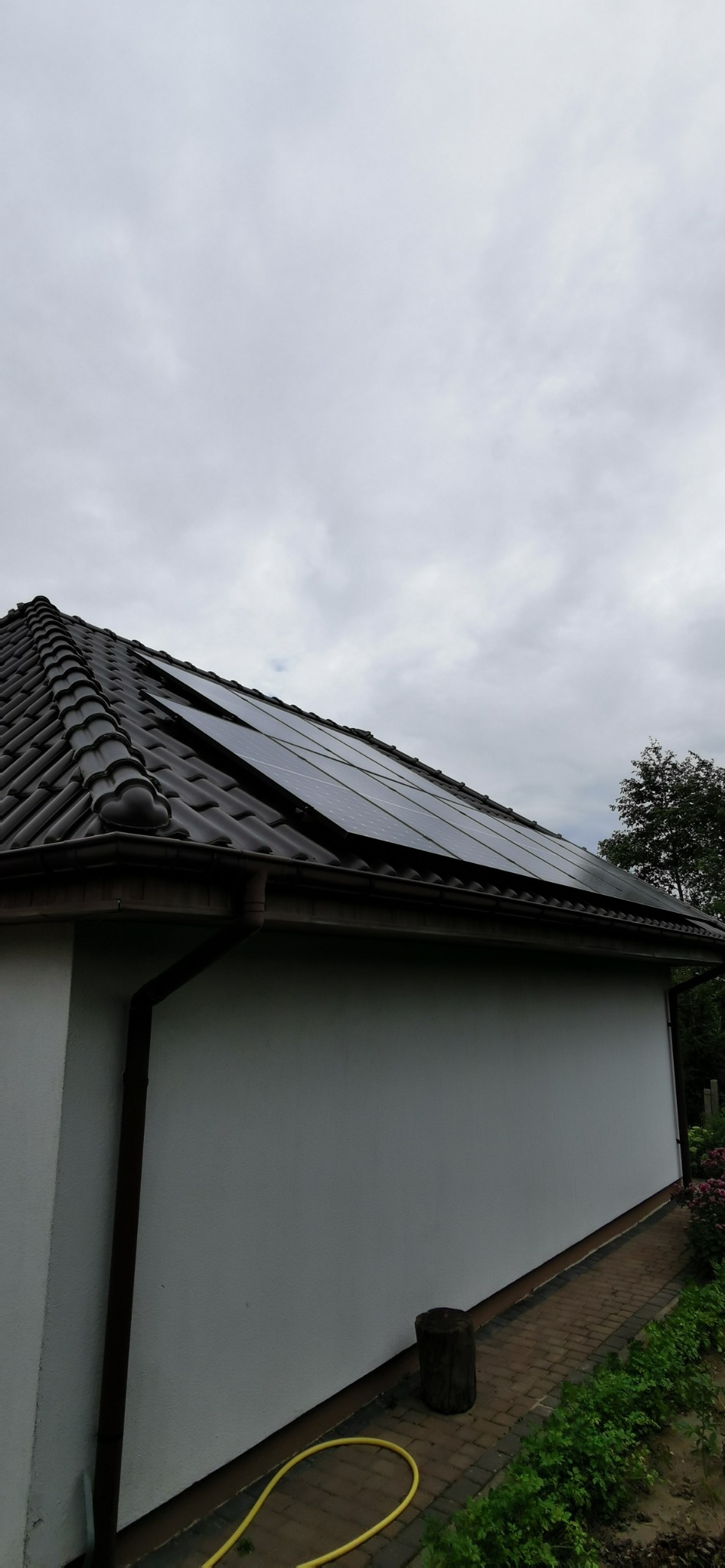 Panele słoneczne na brązowym dachu z dachówki ceramicznej, widok z dołu na białą ścianę budynku w pochmurny dzień.