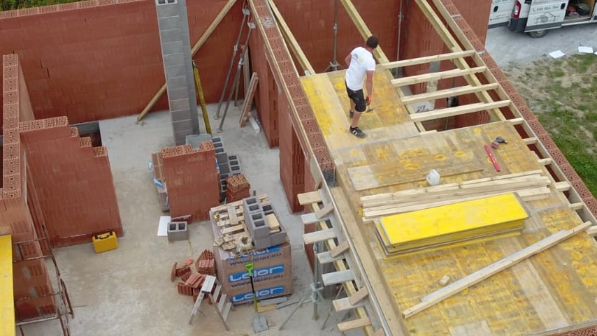 Wznoszenie ścian domu z czerwonej cegły, widok z góry. Na budowie pracownik w białej koszulce. Widoczne materiały budowlane i elementy konstrukcji dachu.