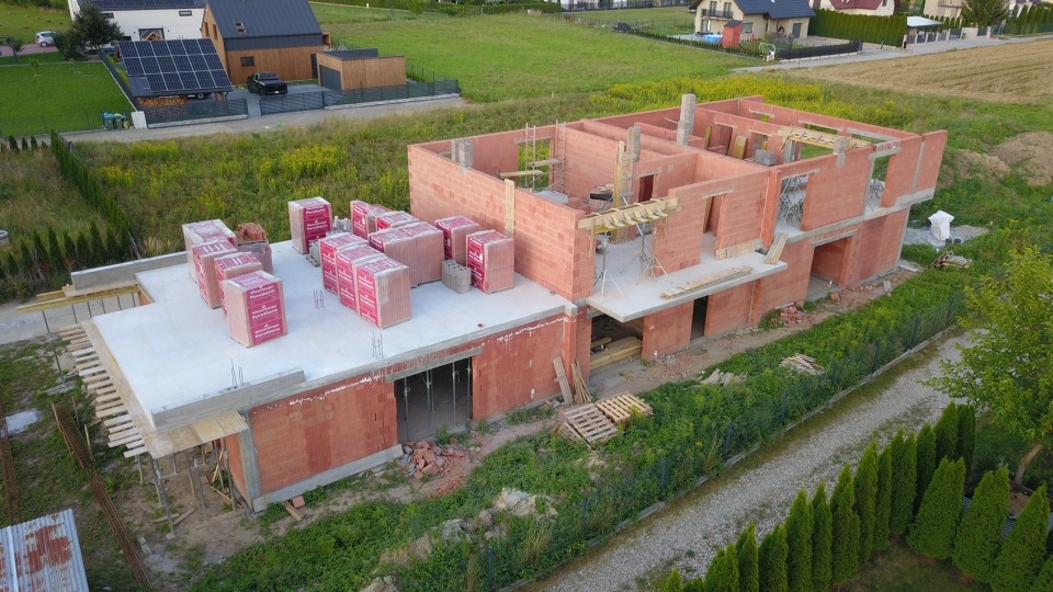 Budowa domu z czerwonej cegły na działce, widok z góry. Na stropie ułożone palety z cegłami. W tle zabudowa jednorodzinna i zieleń. Prace murarskie w toku.