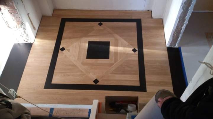 Ułożona podłoga z paneli w geometryczny wzór z czarnymi akcentami, widoczne narzędzia i materiały w tle, częściowo zakryte przez osobę w trakcie pracy.