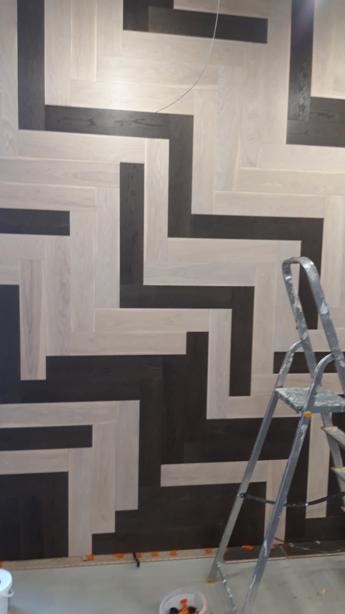 Dwukolorowy parkiet ułożony we wzór jodełki francuskiej, z widocznymi elementami drabiny malarskiej i akcesoriami montażowymi na podłodze.