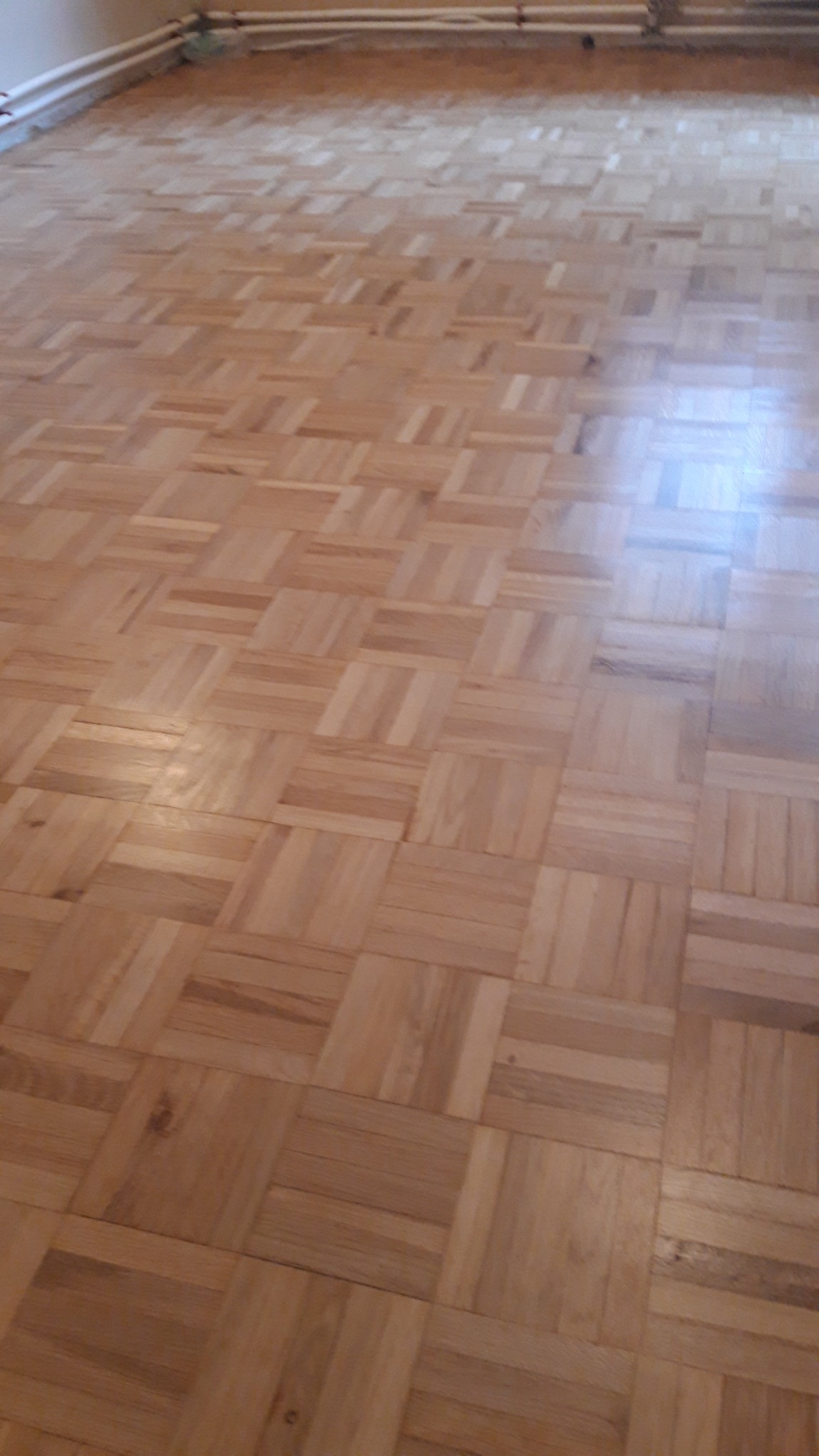 Parkiet ułożony w geometryczny wzór, widoczne ślady po cyklinowaniu na części powierzchni, rury grzewcze przy suficie.