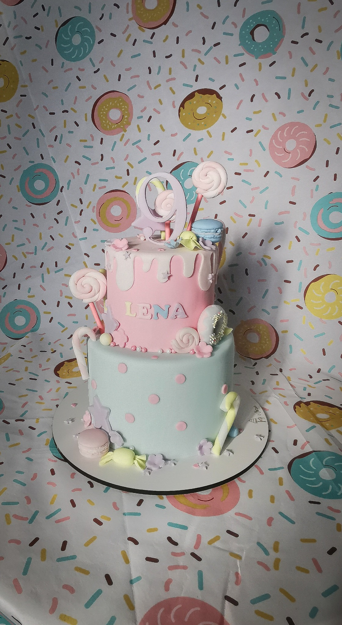 Dwupiętrowy tort w pastelowych kolorach, udekorowany lizakami, makaronikami i napisem 'Lena', na tle wzorzystej tkaniny z motywem pączków i posypki.