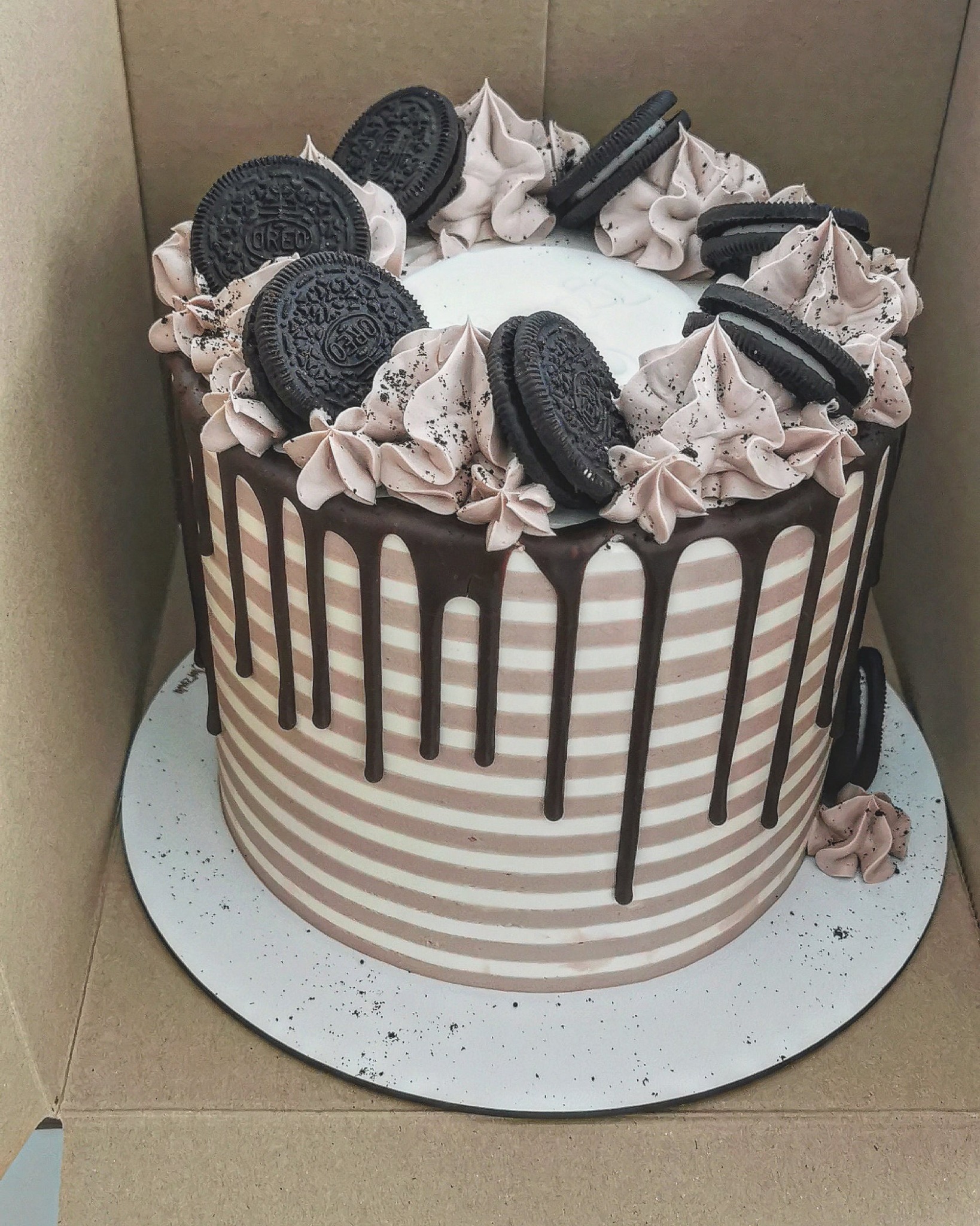 Wysoki tort w paski, z polewą czekoladową spływającą po bokach, udekorowany kremem w kolorze kawy i ciasteczkami Oreo, umieszczony w kartonowym pudełku.