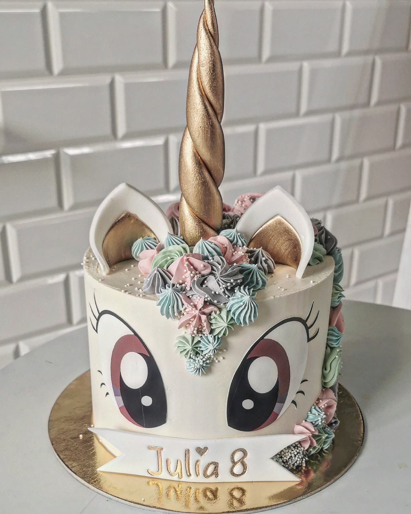 Tort urodzinowy w kształcie jednorożca dla Julii, z kremowymi zdobieniami w pastelowych kolorach, złotym rogiem i napisem na wstążce, umieszczony na złotym podkładzie.