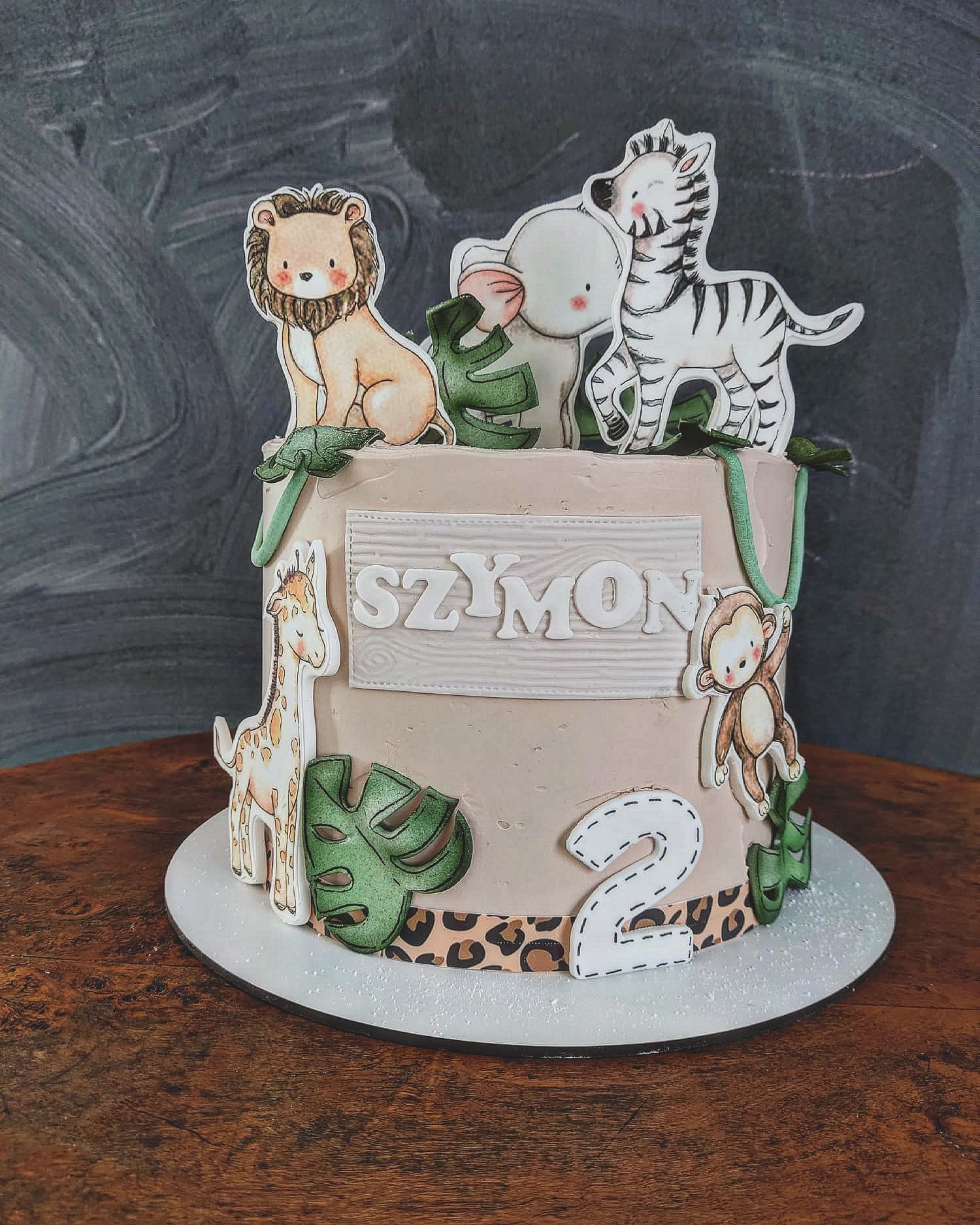 Tort urodzinowy dla dziecka z motywem safari: lew, zebra, słoń, żyrafa, małpka i liście monstery, dekorowany imieniem Szymon i cyfrą 2, na drewnianym stole.