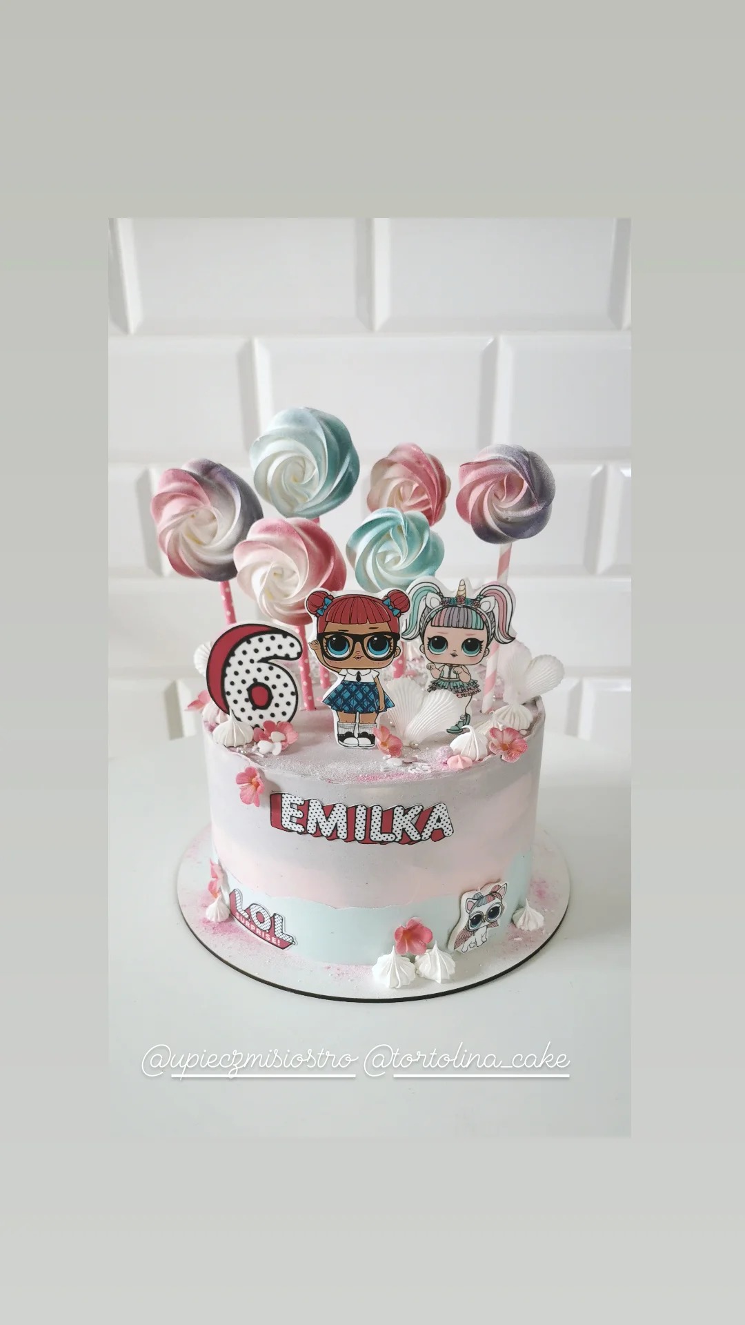 Tort urodzinowy w pastelowych kolorach, zdobiony lizakami bezowymi, postaciami LOL Surprise i imieniem 'Emilka', na białym tle.