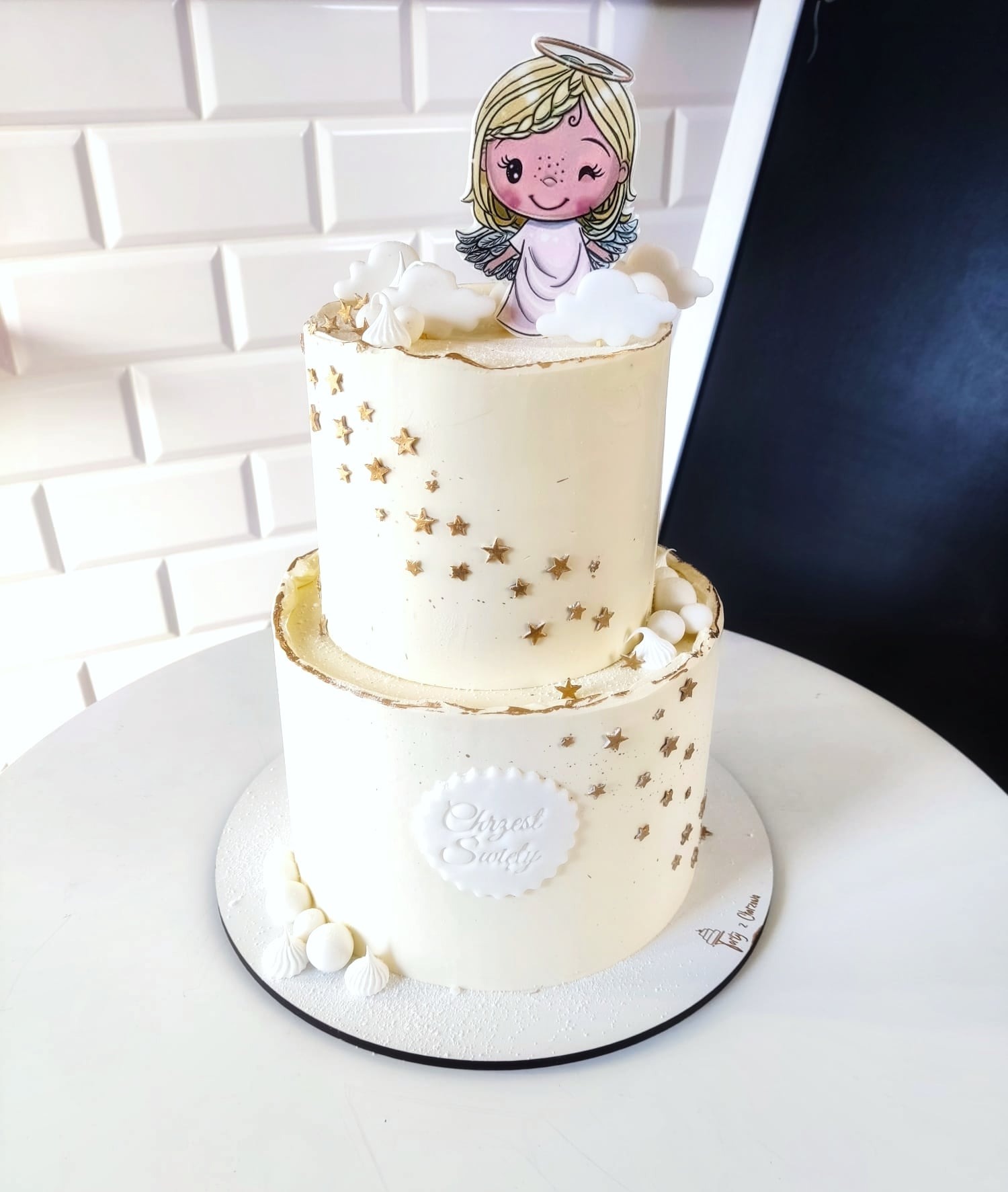 Dwupiętrowy tort z białym kremem, złotymi gwiazdkami, dekoracjami z bezy i papierową ozdobą aniołka z aureolą na wierzchu, umieszczony na białym stole.