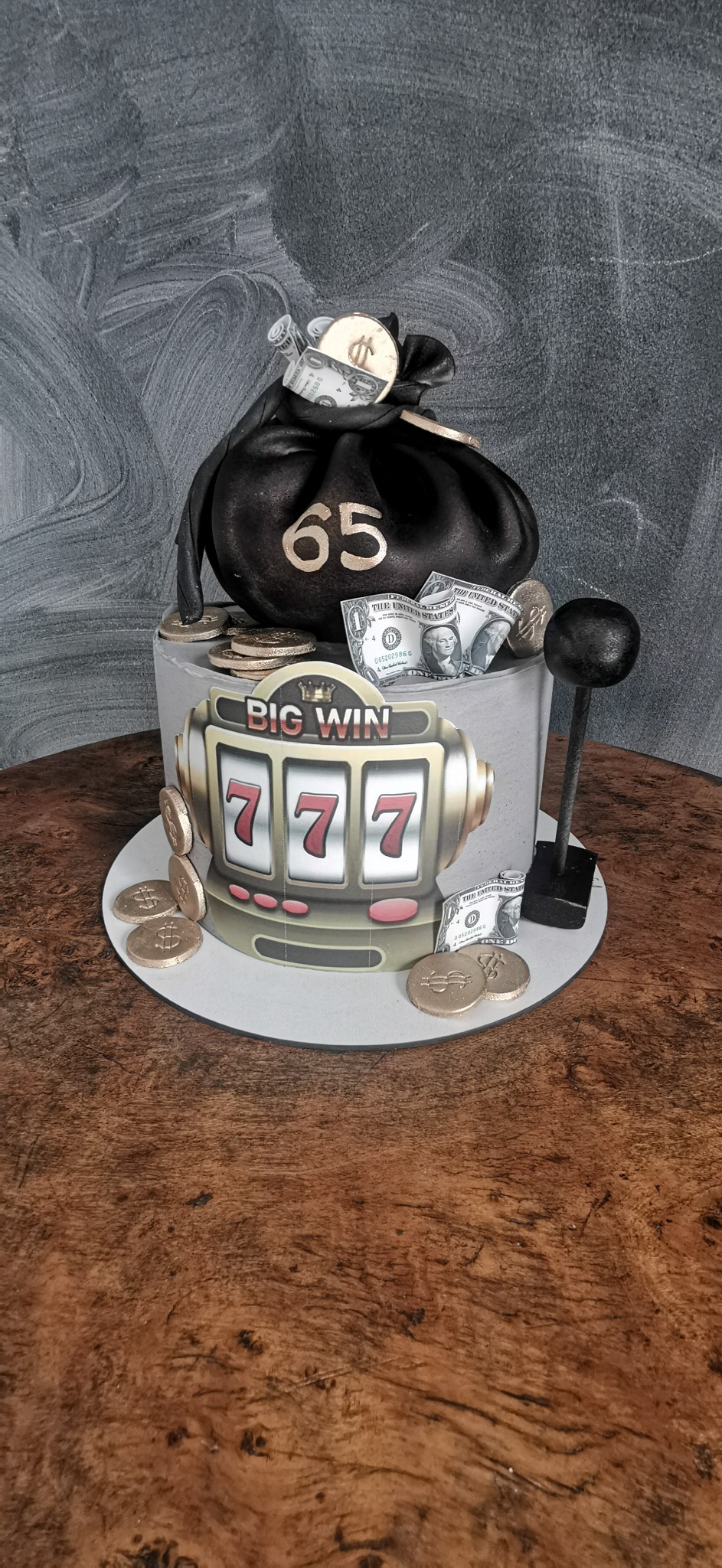 Tort urodzinowy z motywem kasyna: automat z trzema siódemkami i napisem 'Big Win', czarny worek z napisem '65' wypełniony złotymi monetami i zwiniętymi banknotami dolarowymi, całość na drewnianym...
