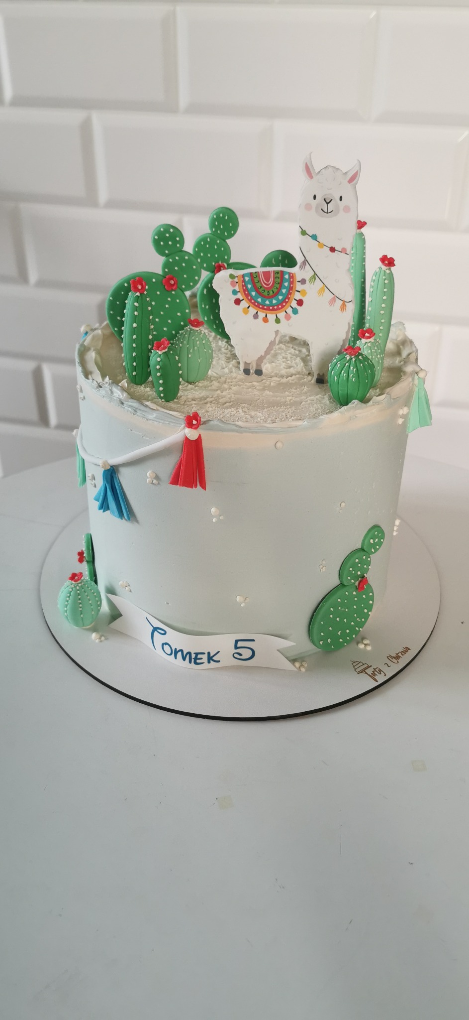 Tort urodzinowy dla dziecka z motywem lamy i kaktusów, dekorowany lukrem i cukrowymi ozdobami, z napisem 'Tomek 5' na białym tle.