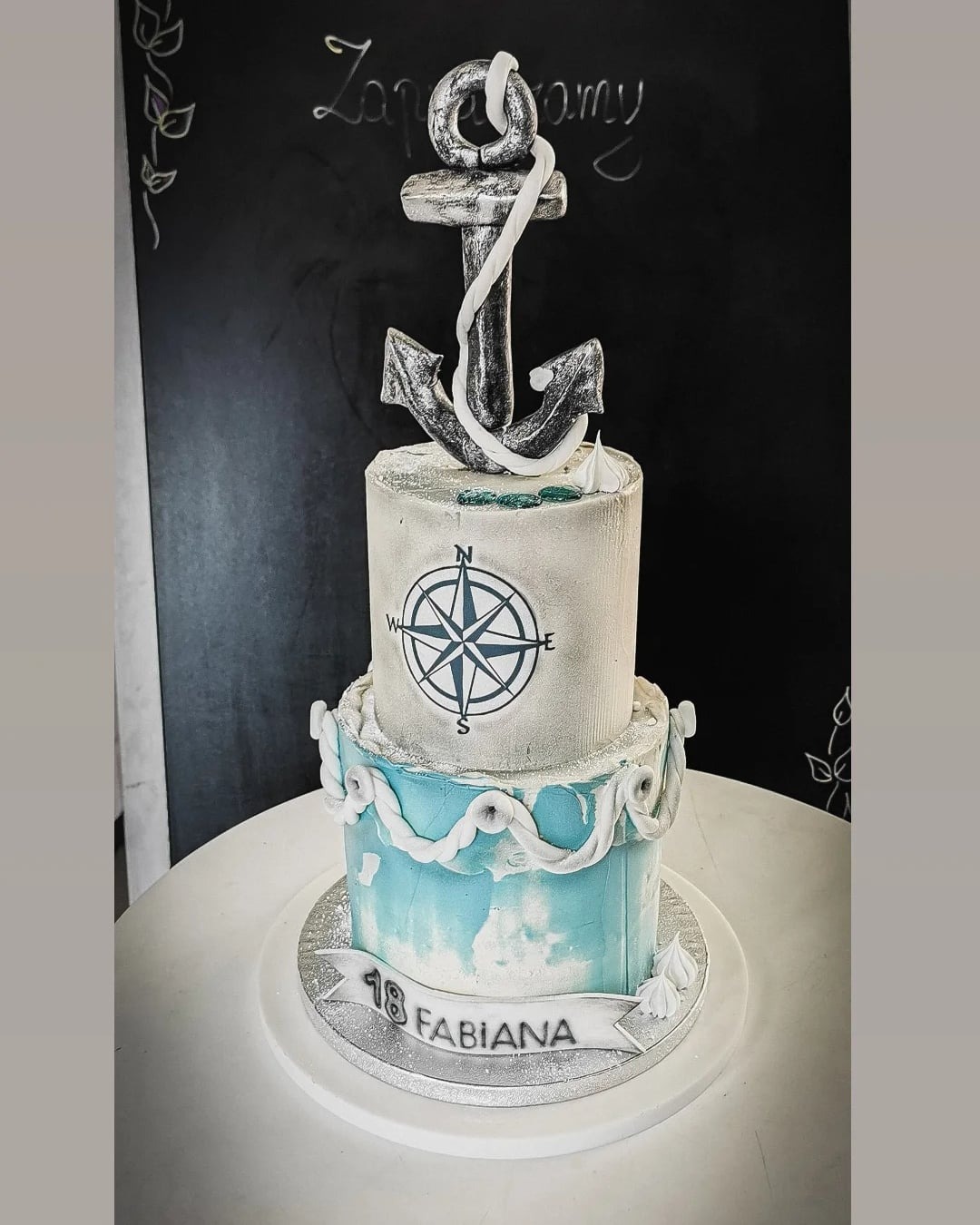 Dwupoziomowy tort z motywem morskim, zdobiony srebrnym kotwicą, kompasem i napisem '18 FABIANA' na tle czarnej tablicy z dekoracjami.