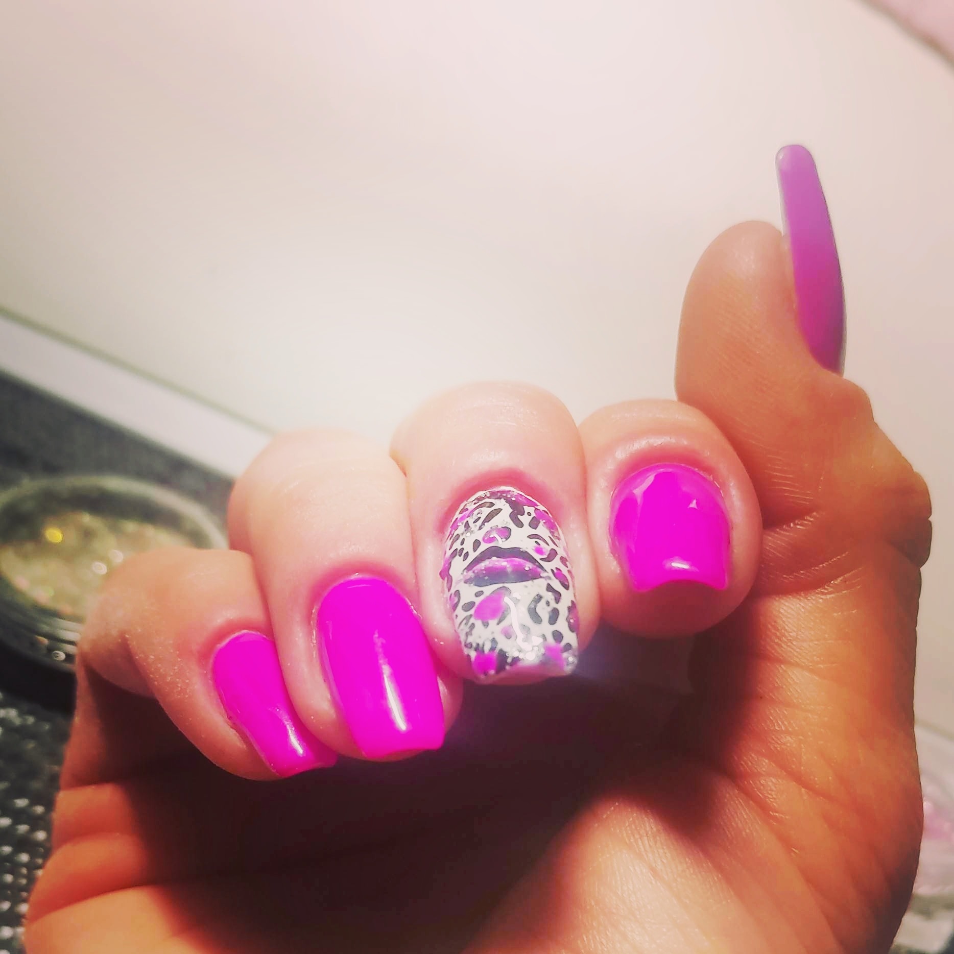 Dłoń z pomalowanymi na różowo paznokciami, jeden z nich z wzorem panterki i ust, na tle rozmytego słoiczka z brokatem.
