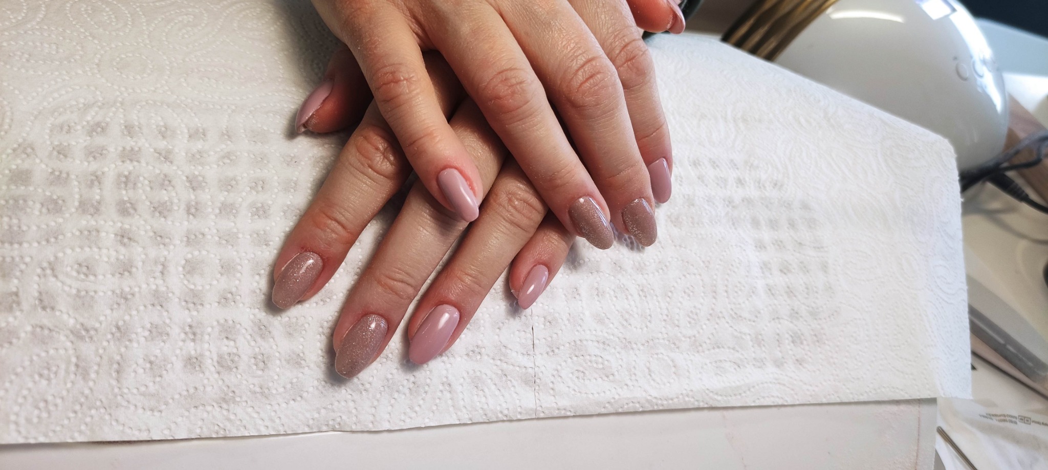 Dłonie z różnymi odcieniami lakieru, jeden z brokatem, na tle białej serwetki i lampy UV do utwardzania lakieru hybrydowego.