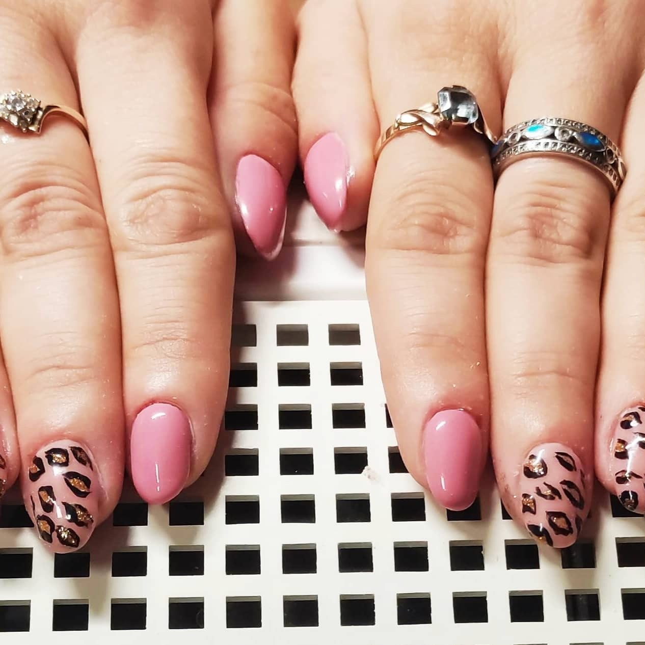 Dłonie z manicure: różowe paznokcie i panterka z brokatem, ozdobione pierścionkami na tle kratki.