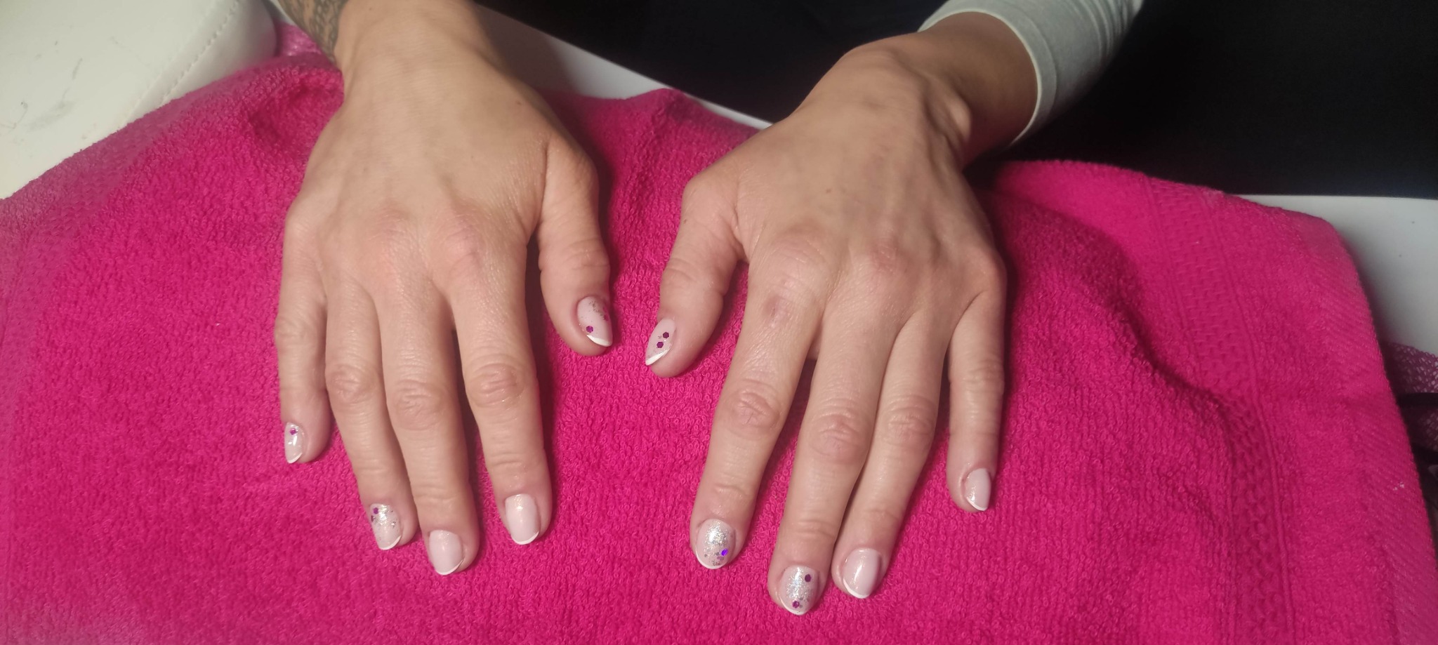 Dłonie po zabiegu upiększającym paznokcie, z białym french manicure i drobnymi, błyszczącymi zdobieniami na różowym ręczniku.