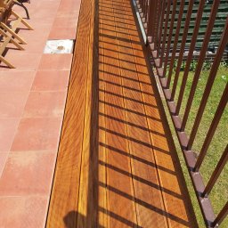 Rafał Różański - Nowa, drewniana podłoga tarasu sąsiaduje z istniejącą, czerwoną terakotą; cień barierki i osoby na deski.