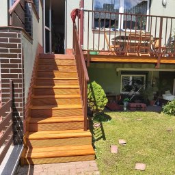 Rafał Różański - Nowe, drewniane schody zewnętrzne z brązową barierką, prowadzące do wejścia w budynku z zieloną elewacją i balkonem, widoczne elementy murarskie z cegły, trawnik z rozrzuconymi płytkami i pędzlem.