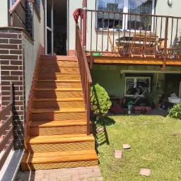 Nowe, drewniane schody zewnętrzne z brązową barierką, prowadzące do wejścia w budynku z zieloną elewacją i balkonem, widoczne elementy murarskie z cegły, trawnik z rozrzuconymi płytkami i pędzlem.