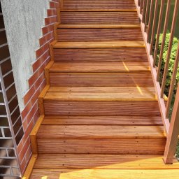 Rafał Różański - Drewniane schody zewnętrzne z ryflowanymi deskami, obramowane czerwoną cegłą, prowadzące w górę, z brązową metalową barierką po prawej stronie.