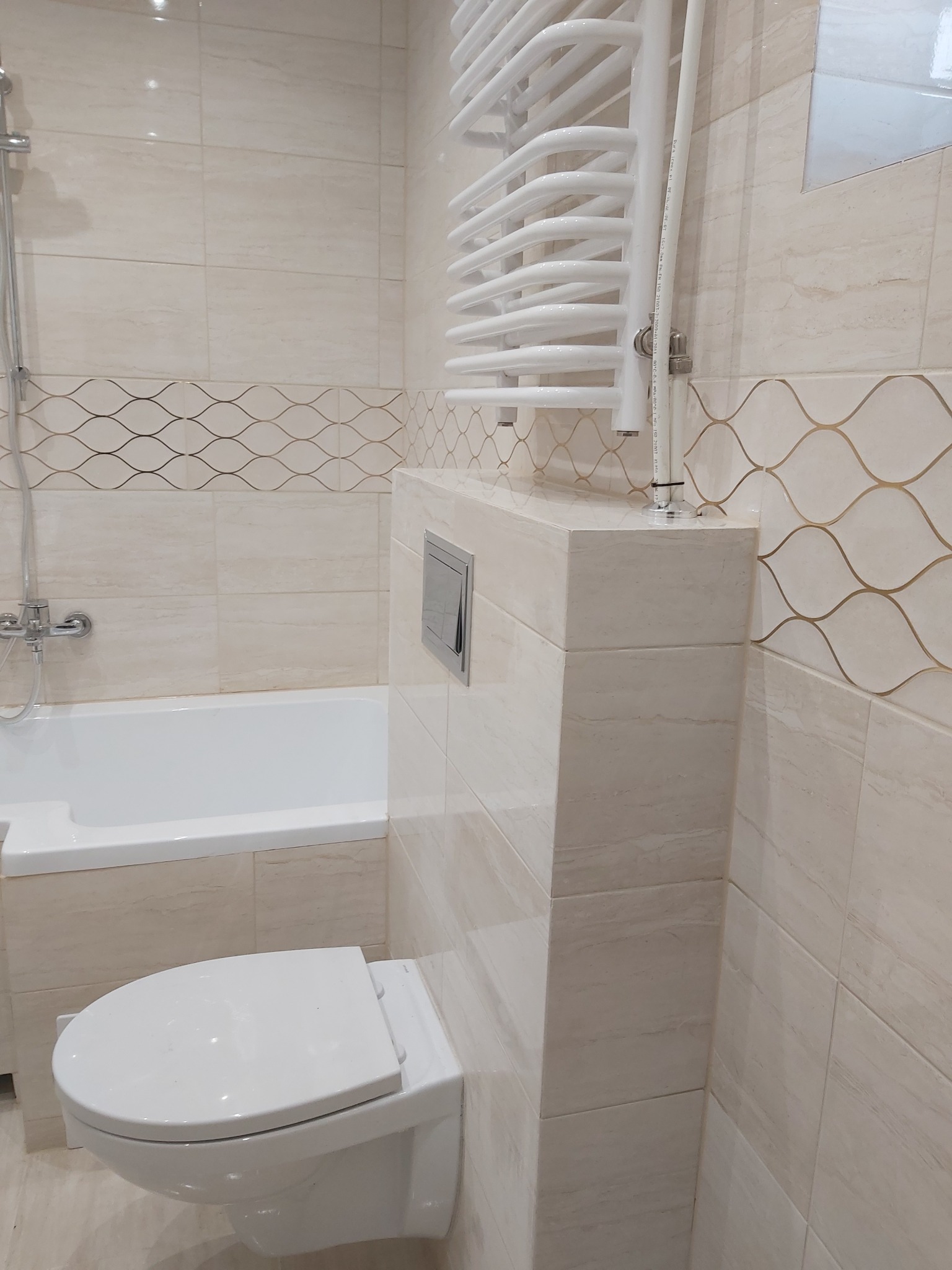 Jasna łazienka z beżowymi płytkami imitującymi trawertyn, dekoracyjnym pasem płytek z motywem fali, podwieszaną toaletą, grzejnikiem drabinkowym i wanną z prysznicem.