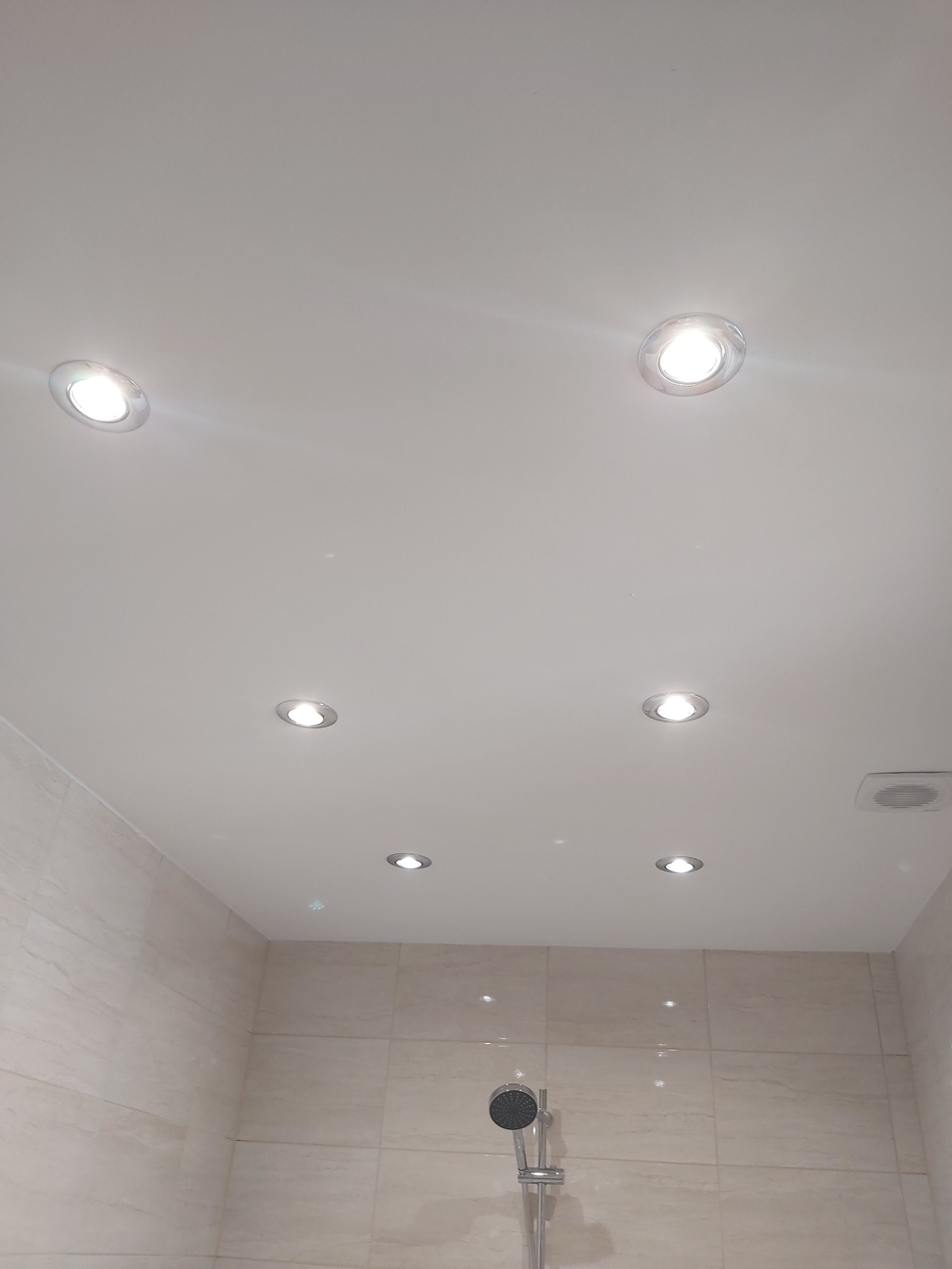 Biały sufit z zamontowanymi sześcioma punktowymi lampami LED w łazience wyłożonej beżowymi płytkami, widoczny fragment prysznica.
