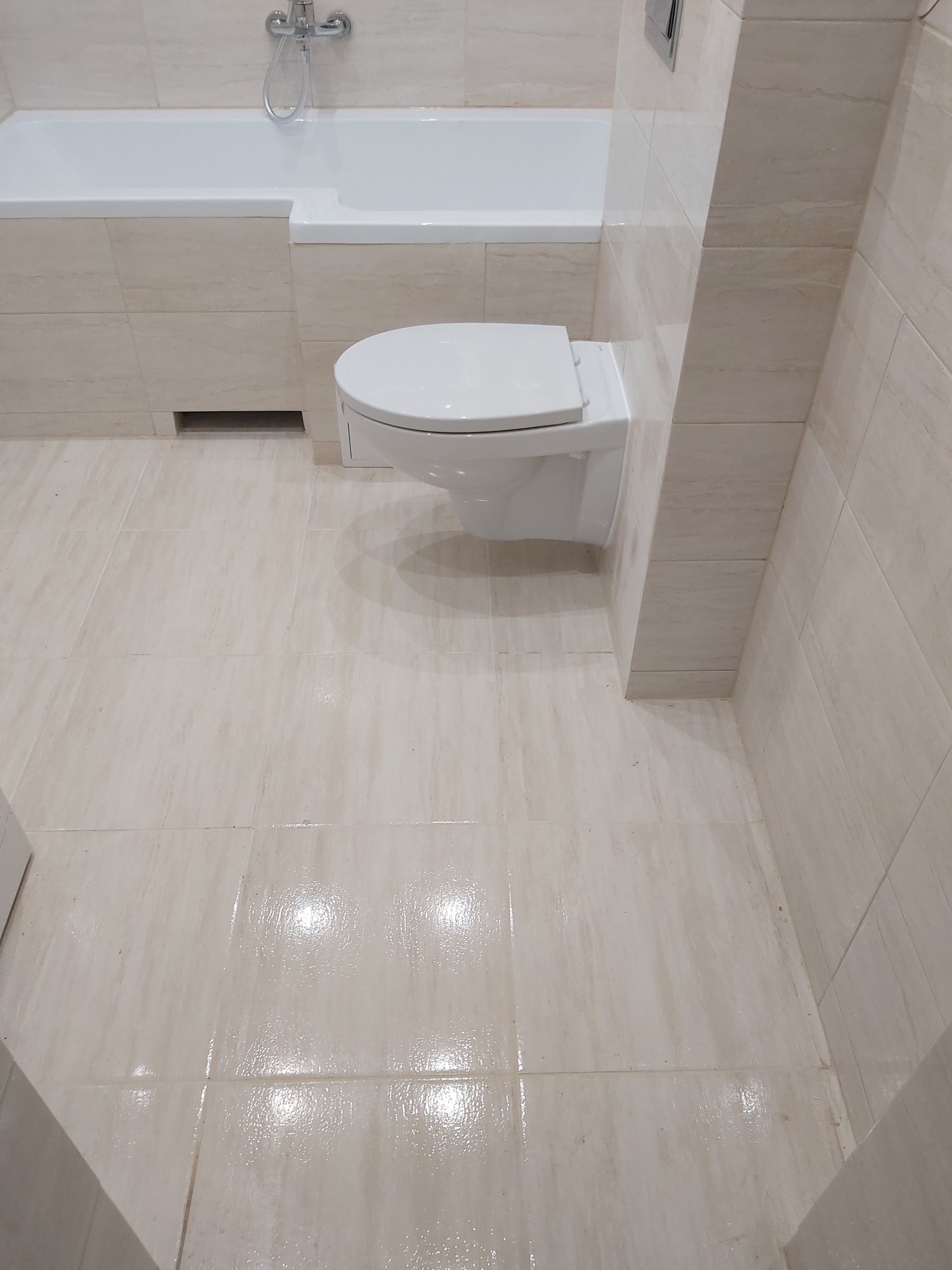 Wykończona łazienka z beżowymi płytkami imitującymi marmur, wisząca toaleta i wanna z chromowaną armaturą.