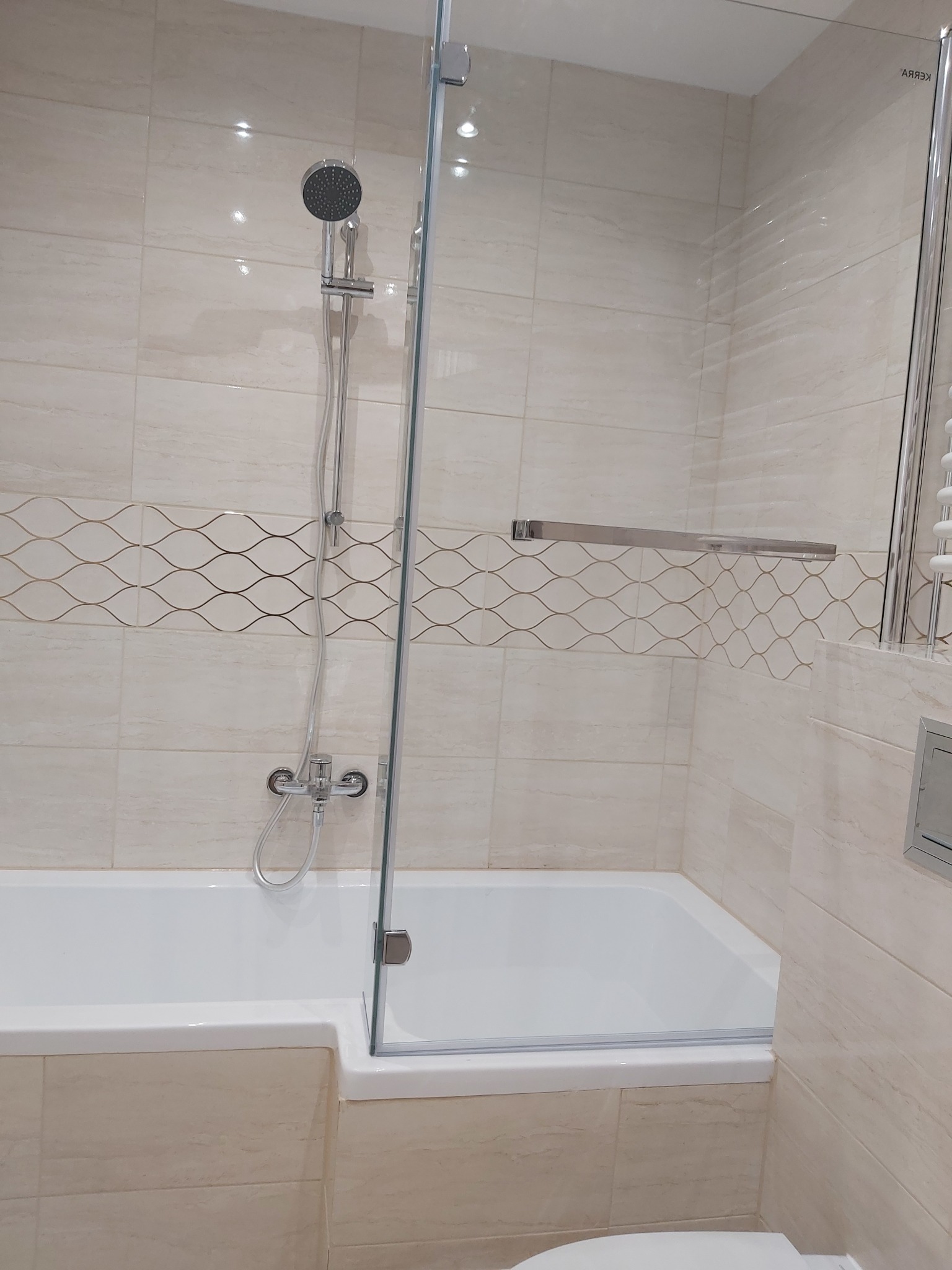 Wykończona łazienka z beżowymi płytkami i dekoracyjnym wzorem, biała wanna z szklanym parawanem, chromowana bateria i deszczownica.