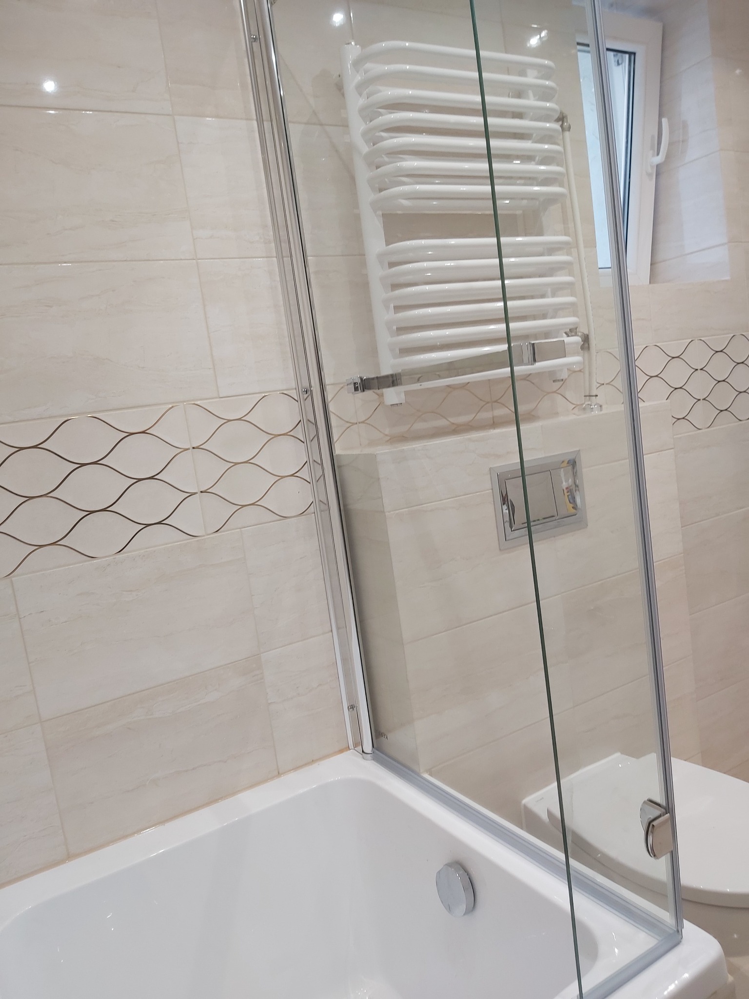 Jasna łazienka po remoncie z wanną, szklanym parawanem nawannowym, grzejnikiem drabinkowym i beżowymi płytkami z dekoracyjnym pasem z motywem fal.