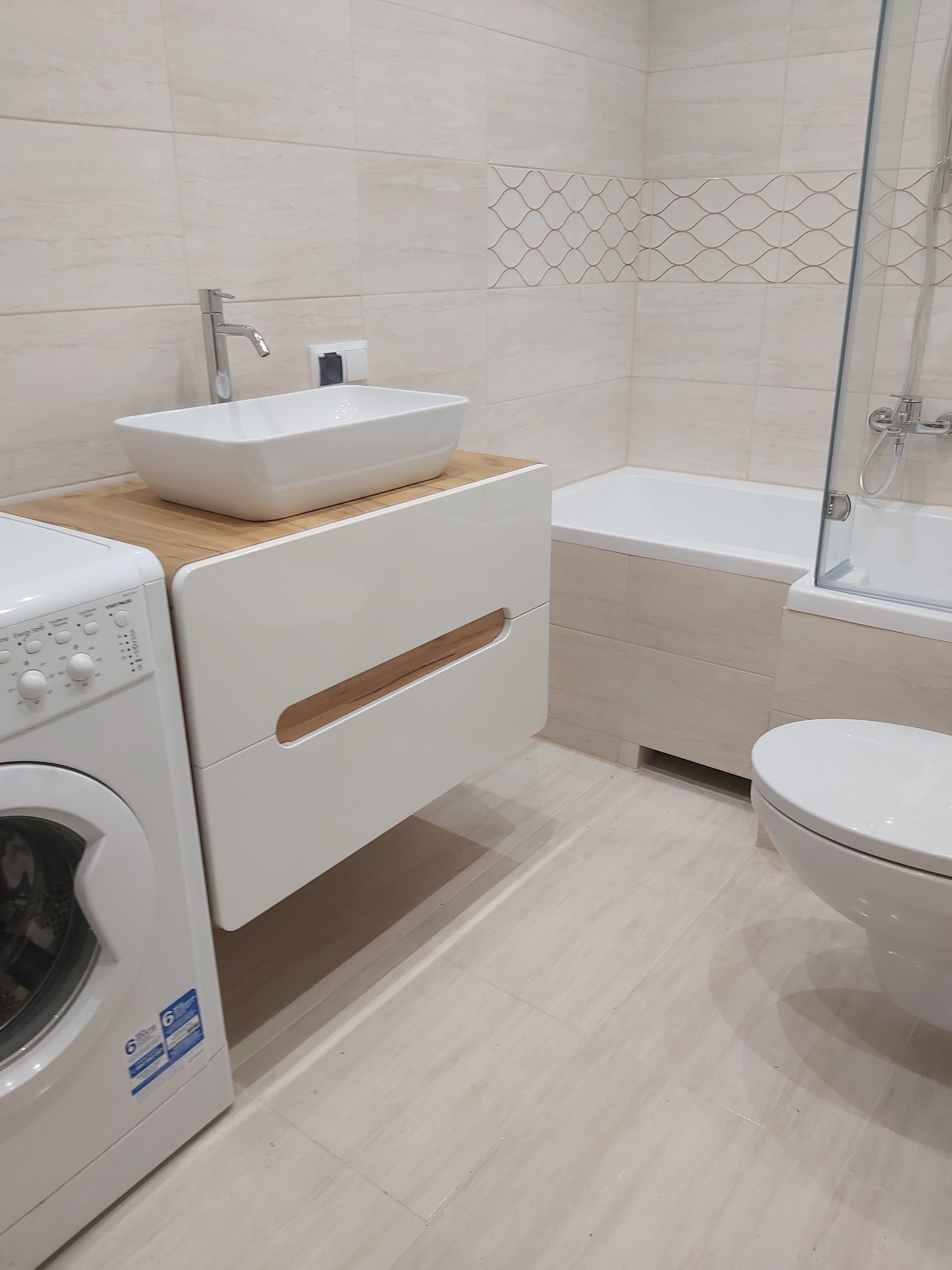 Jasna łazienka z beżowymi płytkami, białą armaturą, szafką pod umywalką z drewnianym blatem, pralką i kabiną prysznicową.