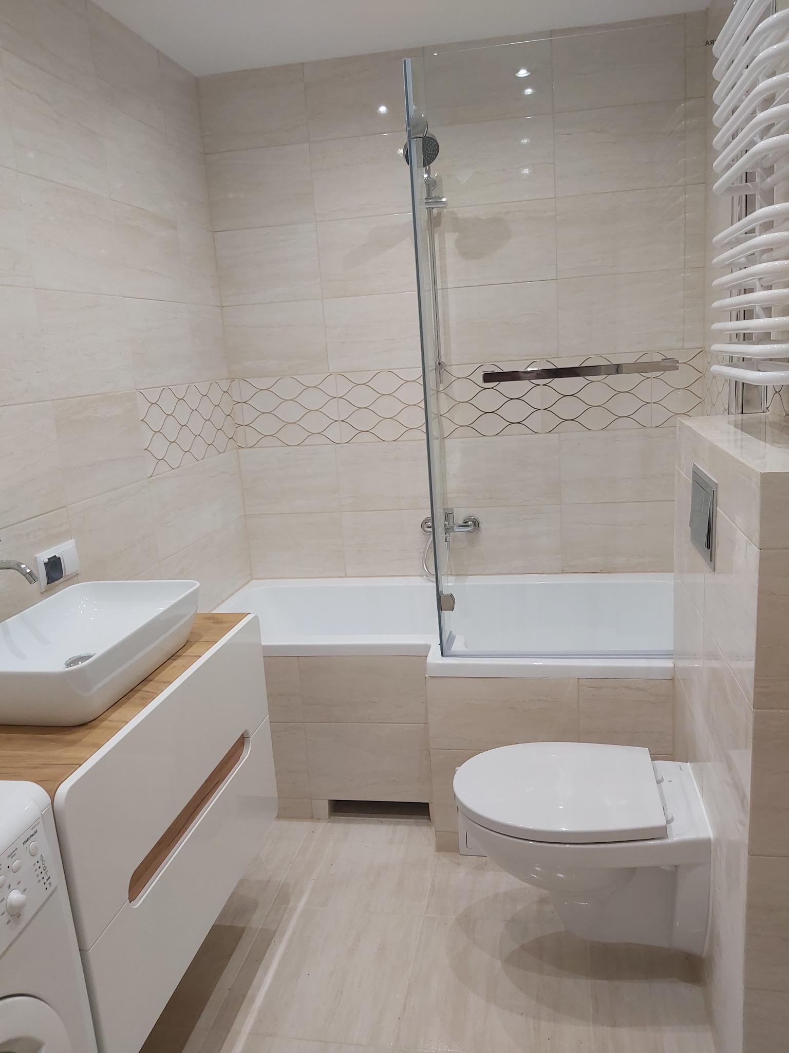 Jasna łazienka z beżowymi płytkami, białą wanną z parawanem, umywalką nablatową na szafce z drewnianym blatem i podwieszaną toaletą.
