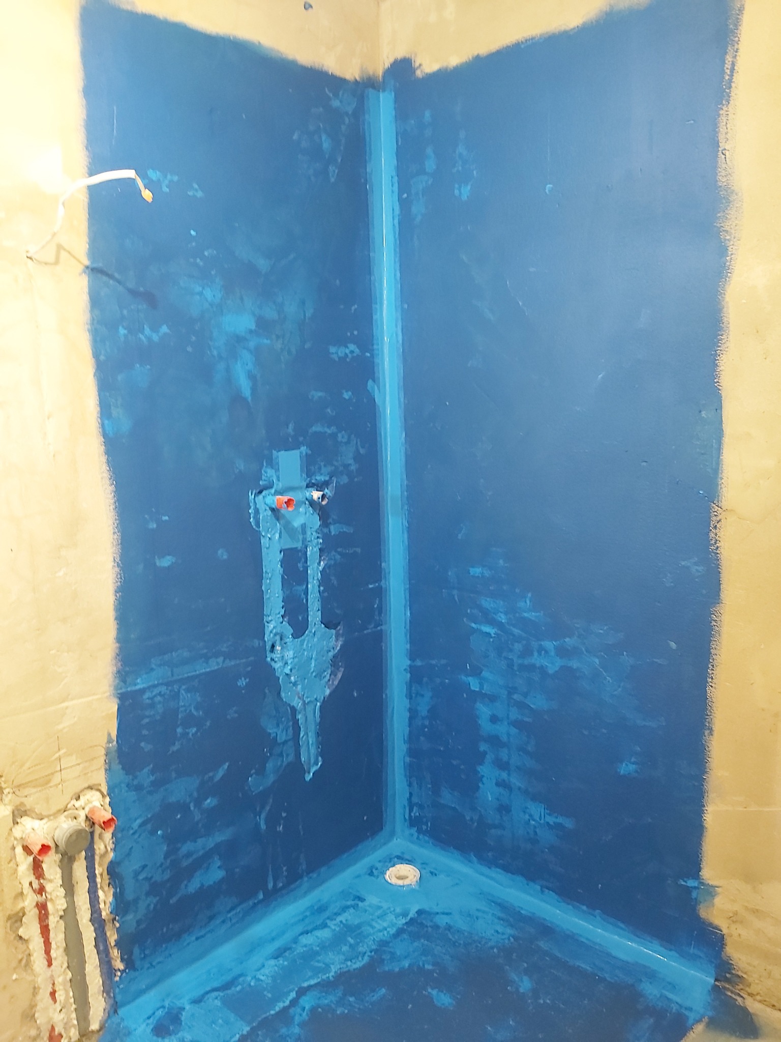 Hydroizolacja w narożnej kabinie prysznicowej, nałożona w kolorze niebieskim, widoczne rury i odpływ podłogowy.