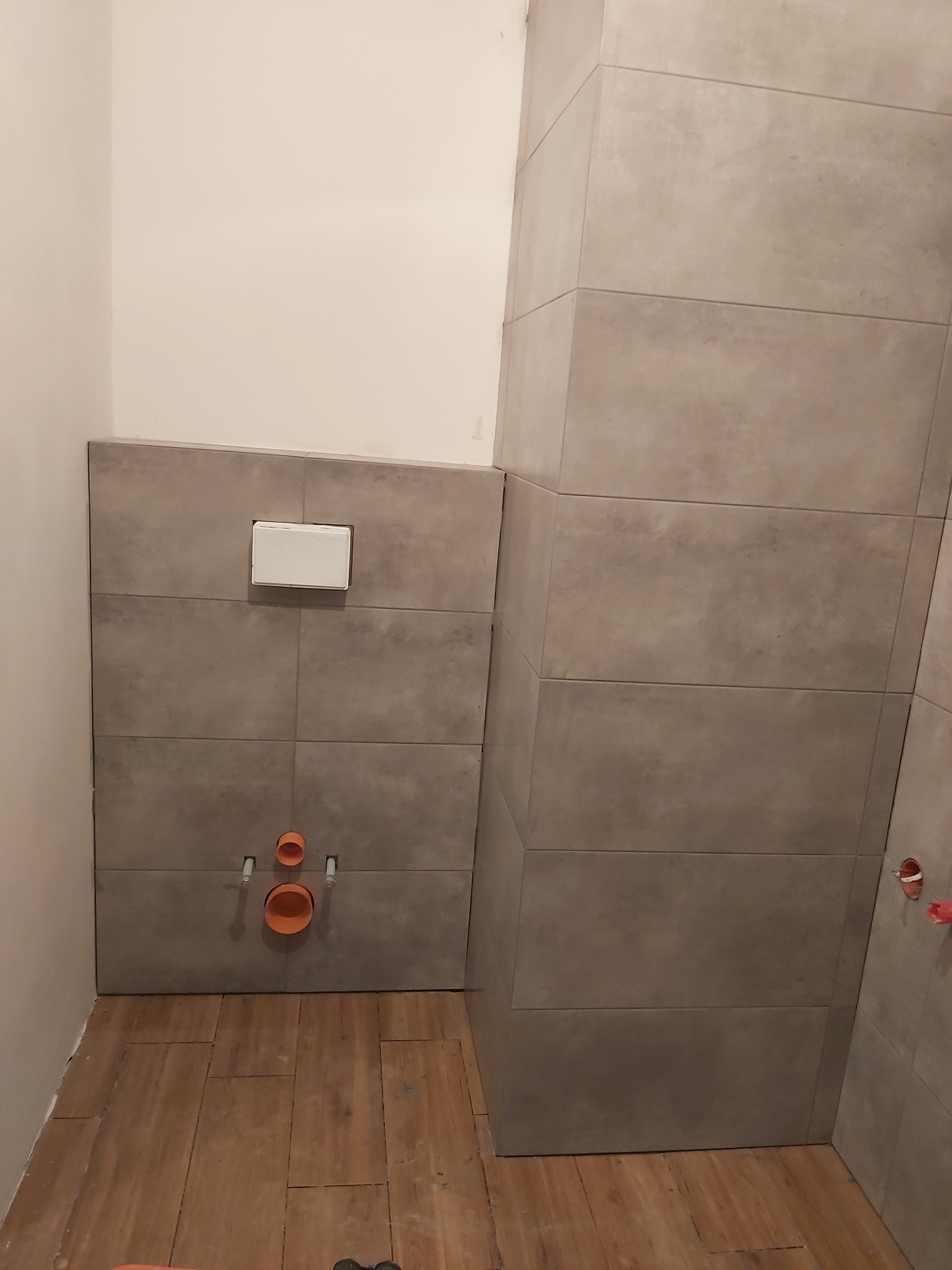 Ściana w łazience wyłożona szarymi płytkami, widoczny podtynkowy stelaż WC z białym przyciskiem oraz pomarańczowe rury kanalizacyjne, na podłodze drewnopodobne płytki.