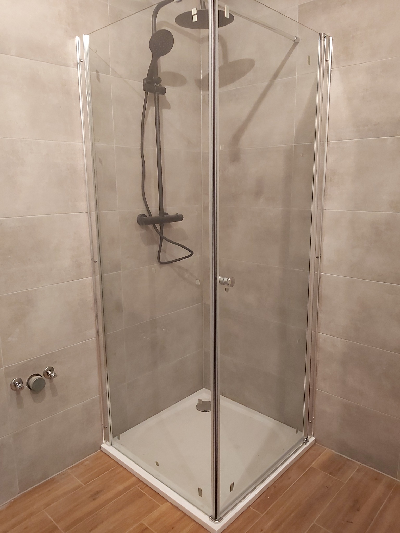 Narożna kabina prysznicowa z czarną armaturą, zamontowana w łazience wyłożonej beżowymi płytkami imitującymi kamień, z drewnopodobną podłogą.