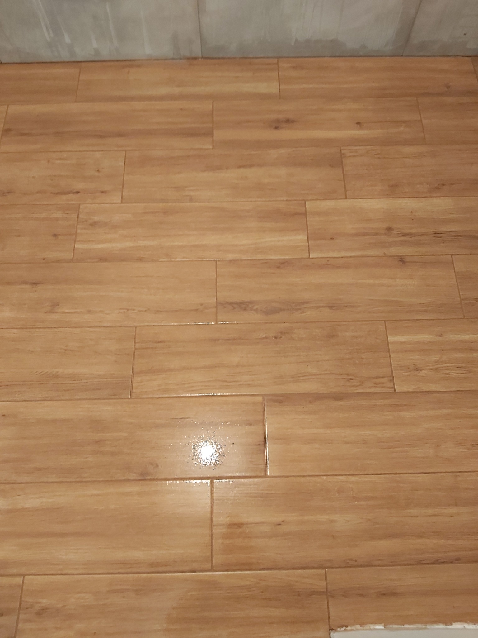 Ułożone podłużne, drewnopodobne płytki podłogowe w odcieniach brązu, z widoczną fugą i fragmentem ściany w tle.