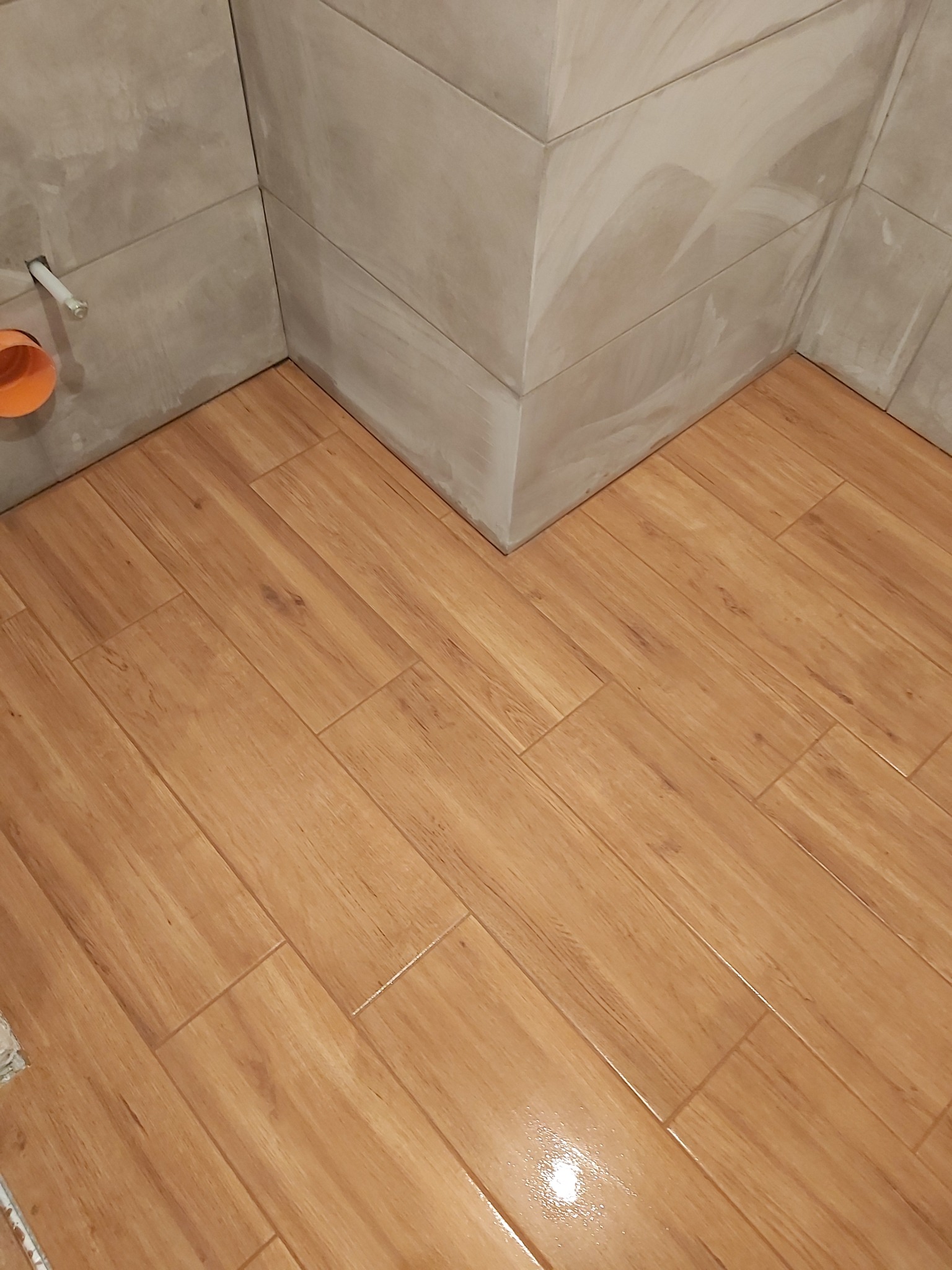 Wykończony róg łazienki z ułożonymi płytkami imitującymi drewno na podłodze i szarymi płytkami na ścianach ułożonymi w jodełkę, widoczny fragment instalacji hydraulicznej.