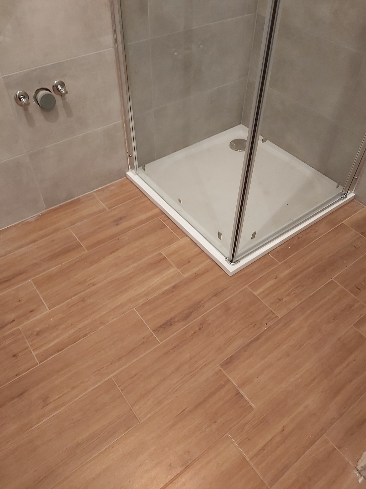 Narożna kabina prysznicowa z białym brodzikiem i szklanymi drzwiami, widoczne trzy chromowane zawory na szarej, kafelkowanej ścianie oraz podłoga wyłożona brązowymi płytkami imitującymi drewno.