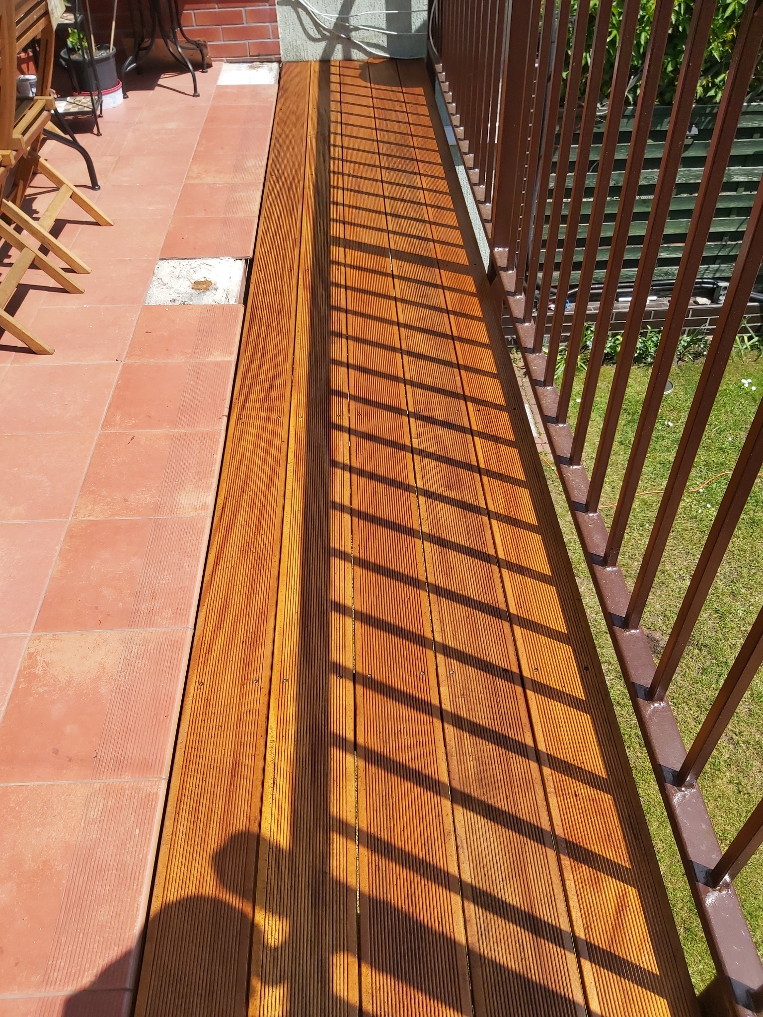 Nowa, drewniana podłoga tarasu sąsiaduje z istniejącą, czerwoną terakotą; cień barierki i osoby na deski.