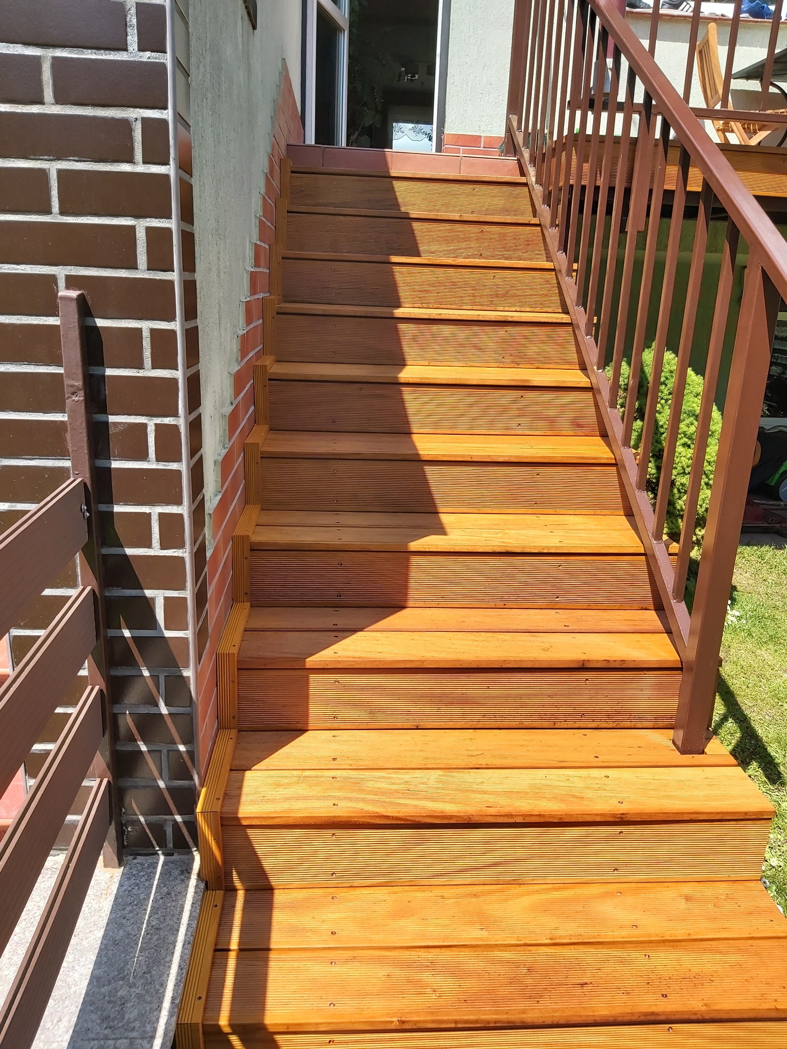 Nowe, drewniane schody zewnętrzne z brązową balustradą przy budynku z cegły w Poznaniu, prowadzące do drzwi wejściowych.