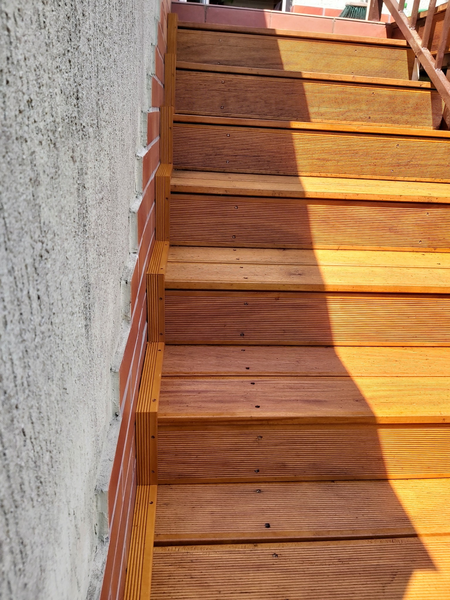 Drewniane schody zewnętrzne z ryflowanymi deskami, przylegające do ściany wykończonej cegłą i szarym tynkiem, oświetlone popołudniowym słońcem.