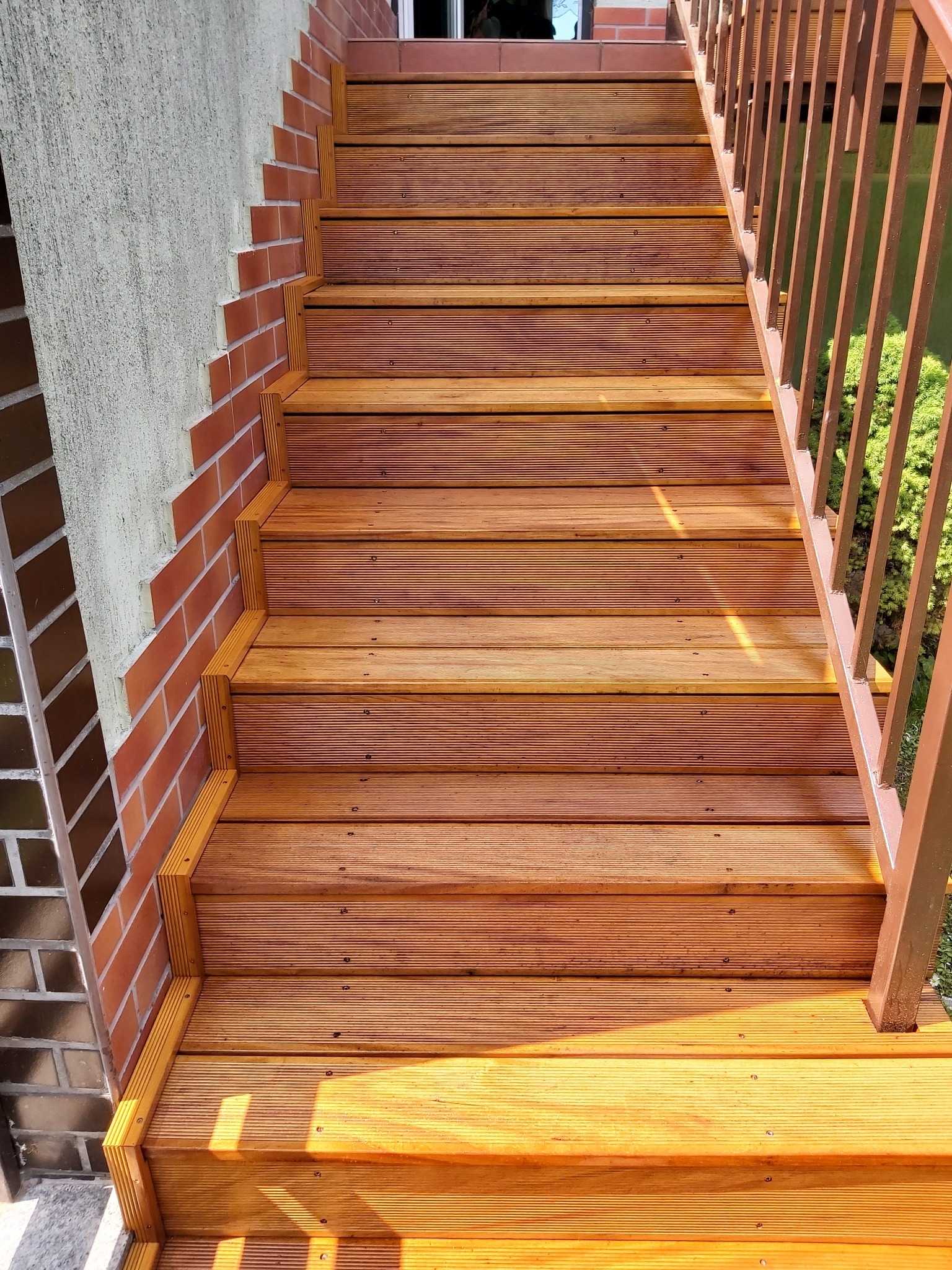 Drewniane schody zewnętrzne z ryflowanymi deskami, obramowane czerwoną cegłą, prowadzące w górę, z brązową metalową barierką po prawej stronie.