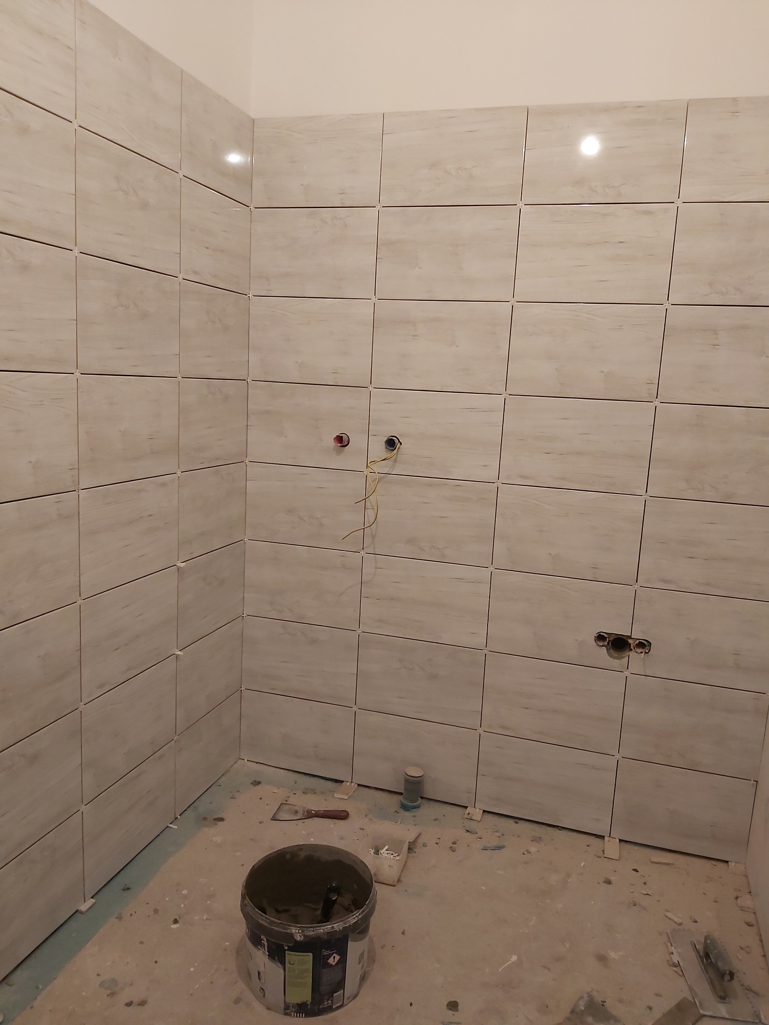 Ściana łazienki w trakcie układania beżowych płytek imitujących drewno, widoczne krzyżyki dystansowe, puszki elektryczne i wiadro z zaprawą, rozmazane tło.