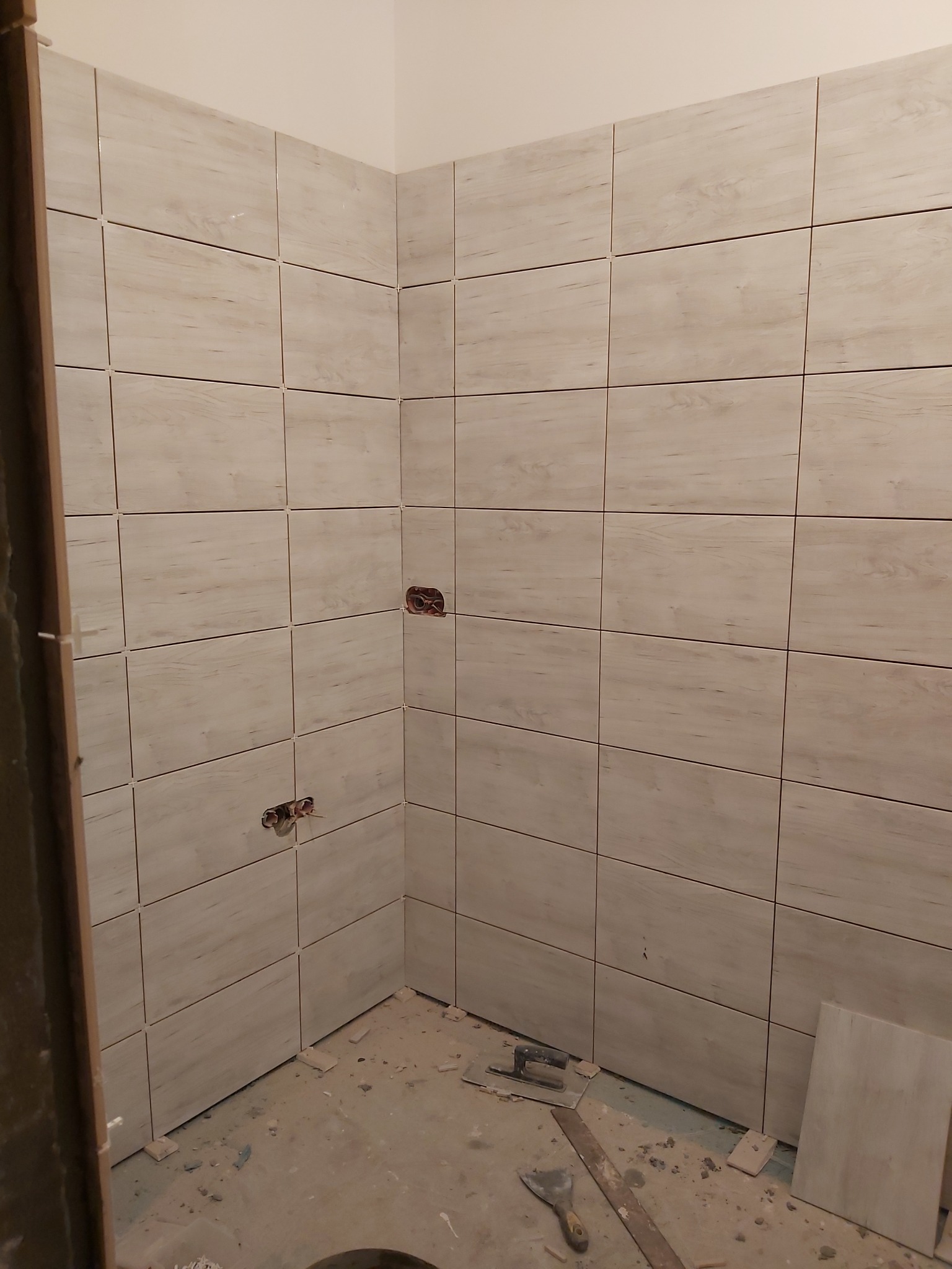 Ściany łazienki wyłożone jasnymi, drewnopodobnymi płytkami ceramicznymi, widoczne otwory na instalacje wodne, na podłodze resztki zaprawy i narzędzia glazurnicze.