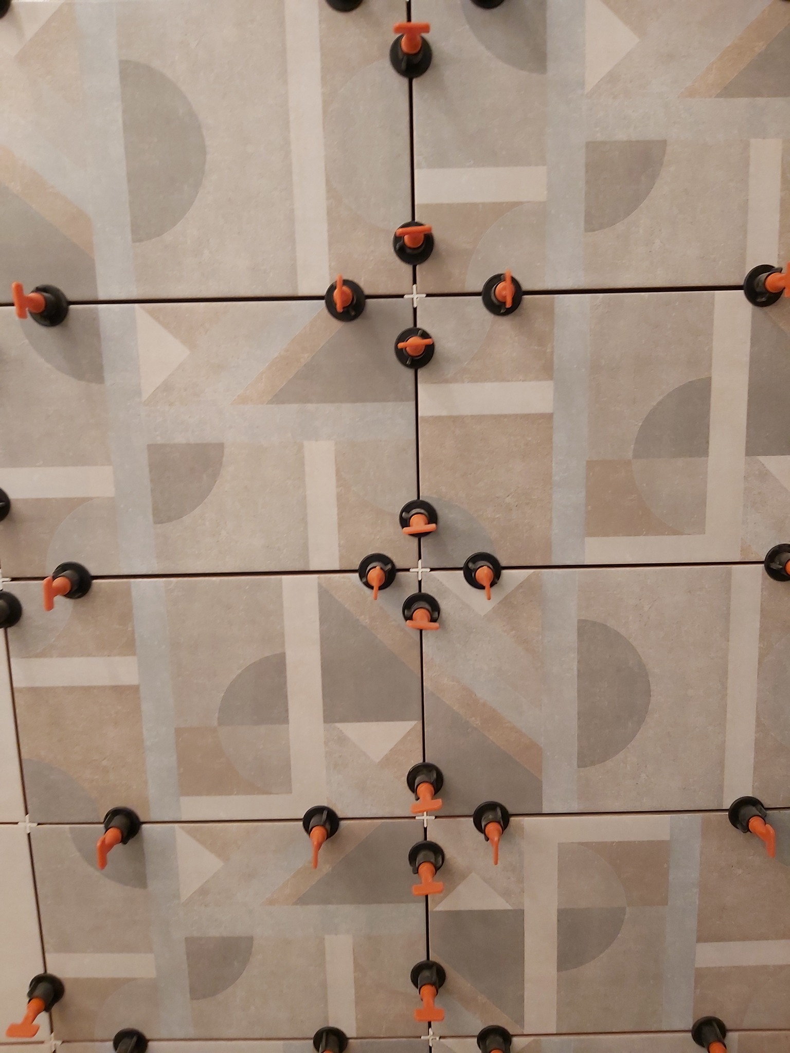 Ułożone płytki ceramiczne z geometrycznym wzorem, zabezpieczone systemem poziomowania z pomarańczowymi klinami i czarnymi nakładkami, widoczne fugi.