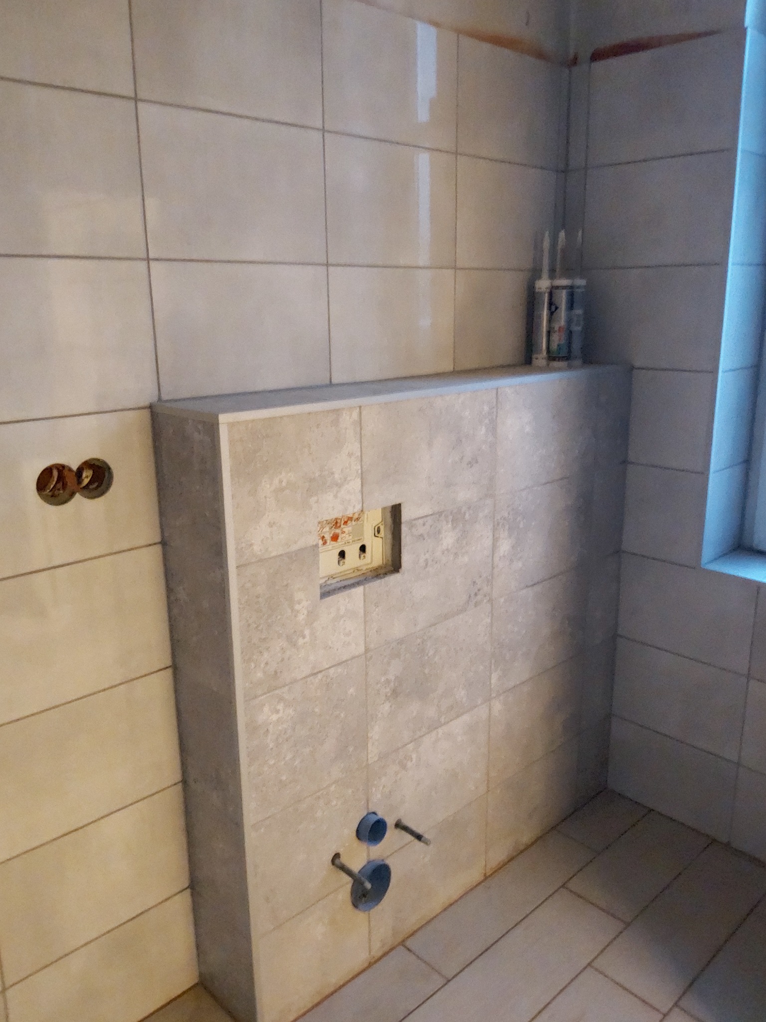 Ściana w łazience z obudową stelaża podtynkowego WC, wykończona jasnymi płytkami ceramicznymi imitującymi beton, widoczne otwory na instalacje wodno-kanalizacyjne oraz puszka elektryczna.