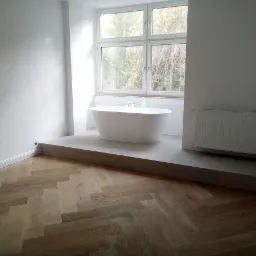 Nowoczesna łazienka z wolnostojącą białą wanną przy oknie, grzejnikiem panelowym na ścianie i parkietem ułożonym w jodełkę.