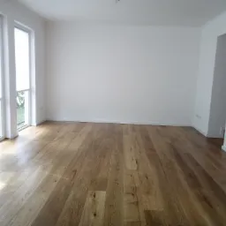 Jasne, drewniane panele podłogowe w pustym, nowo wykończonym pokoju z białymi ścianami i oknami z roletami.