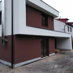 Dwukondygnacyjny dom z brązową i białą elewacją, widoczny stalowy komin i czarna rynna, teren wokół domu w trakcie prac wykończeniowych.
