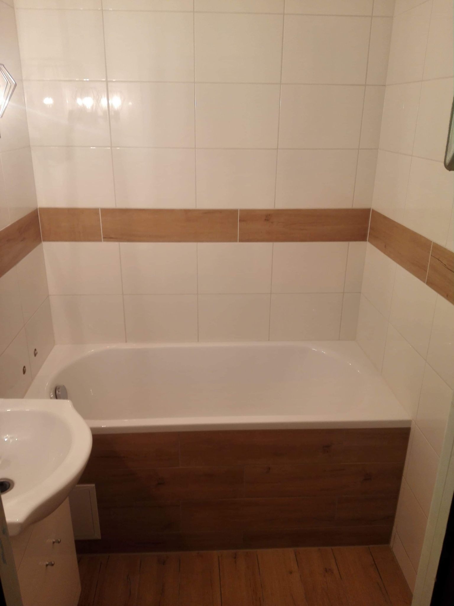 Wykończona łazienka z białą wanną obudowaną panelami imitującymi drewno, białe płytki z poziomym pasem dekoracyjnym w kolorze drewna oraz umywalka na szafce.