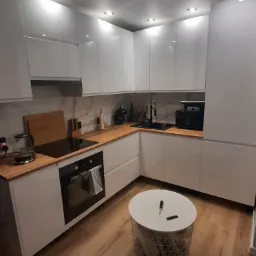 Nowoczesna, biała kuchnia z drewnianymi blatami w Jastrzębiu-Zdroju, oświetlona punktowymi lampami sufitowymi, z czarnym zlewem i sprzętem AGD, uzupełniona okrągłym stolikiem z narzędziami.