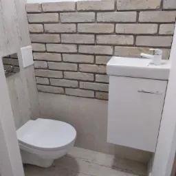 Mała łazienka z toaletą podwieszaną, umywalką nablatową na szafce i ścianą wykończoną dekoracyjną cegłą w odcieniach beżu i szarości.